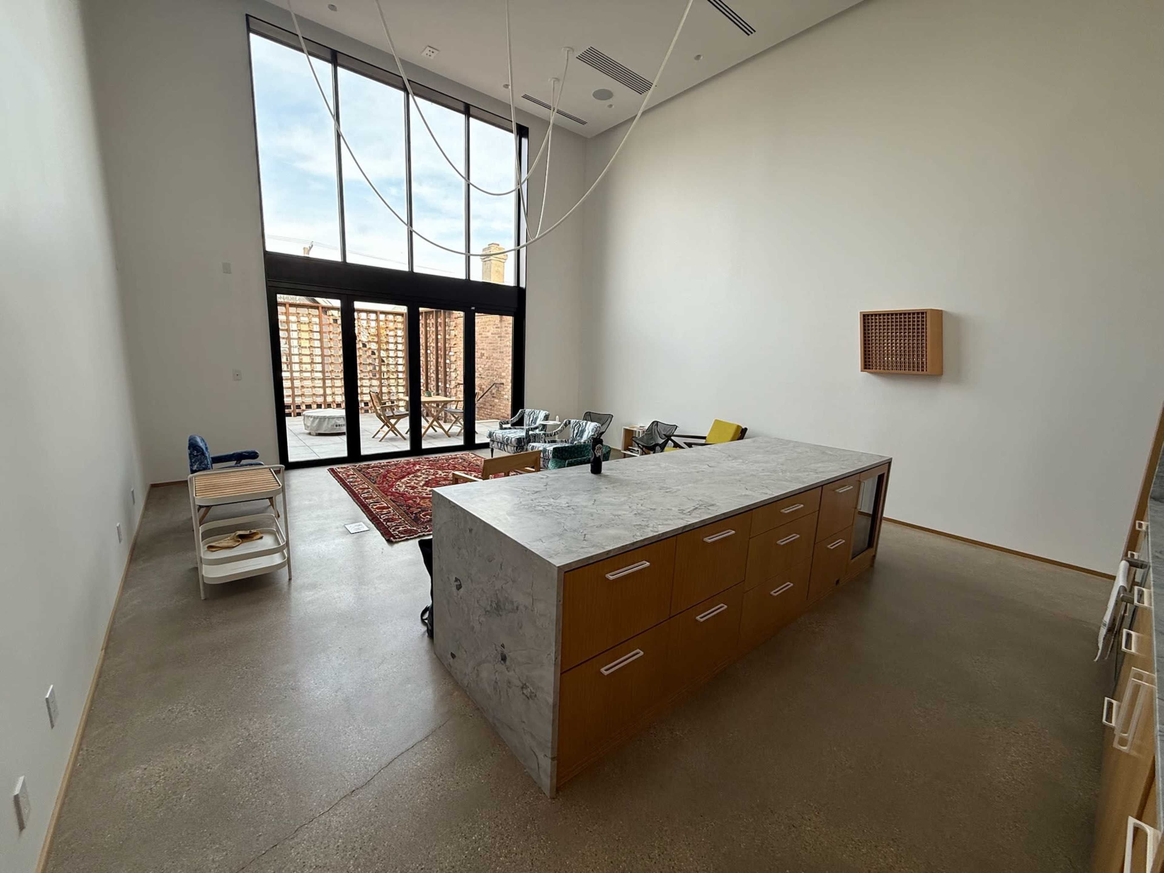 The image shows a spacious, modern interior with a large concrete countertop island, minimal furnishings, and large windows allowing natural light to enter.
