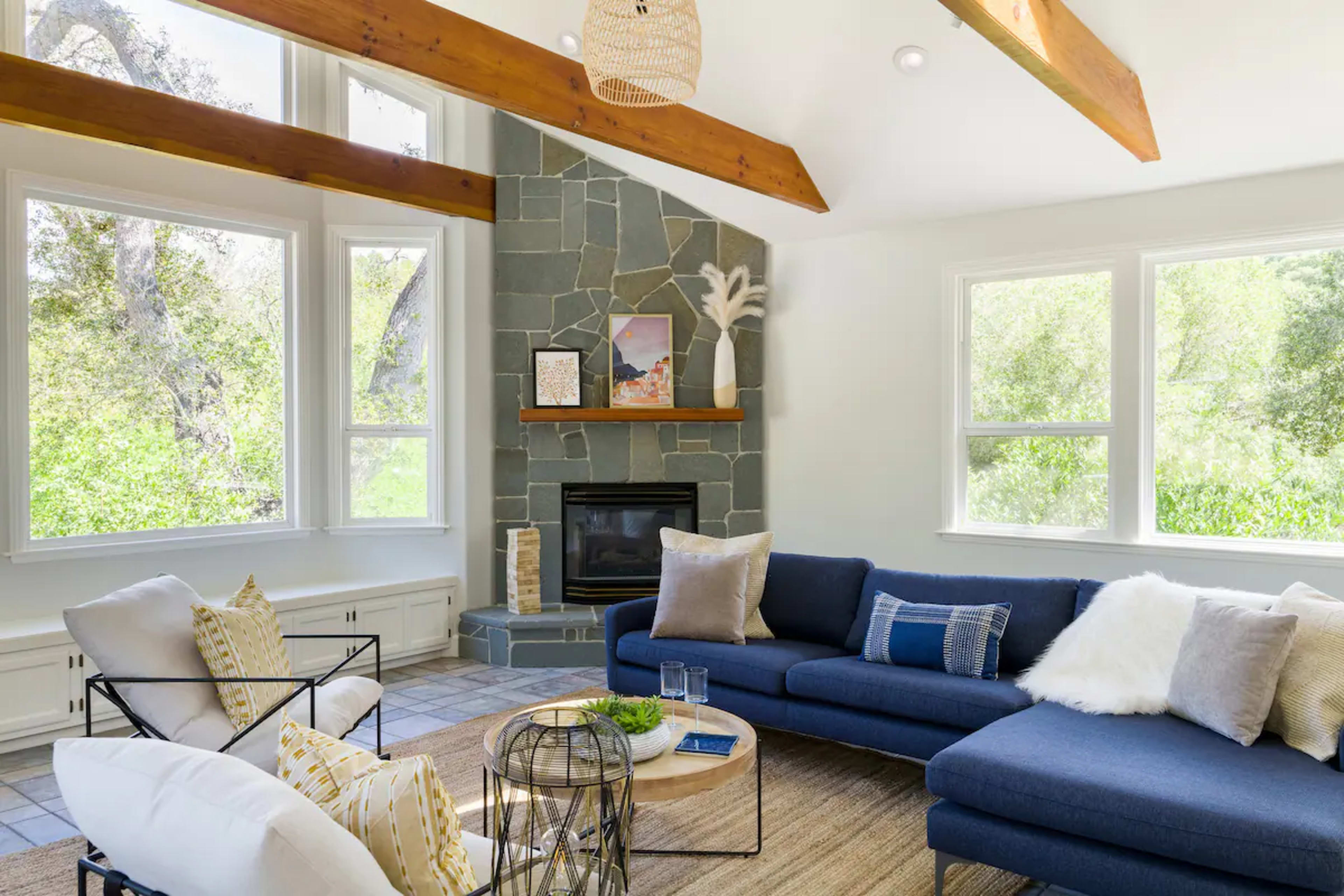 The cozy living room features a blue sectional sofa, a stone fireplace, and large windows with views of greenery.