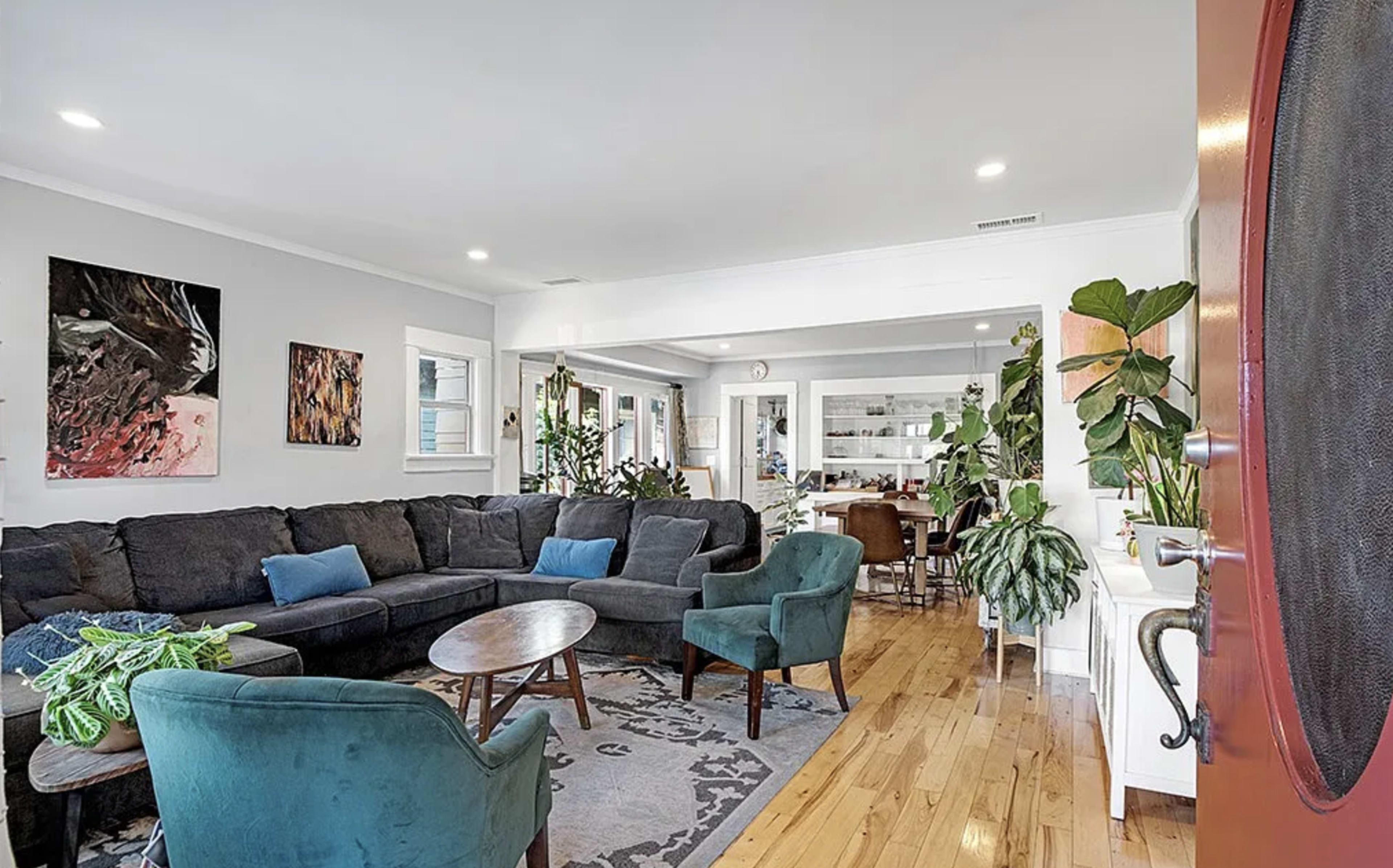 The image shows a spacious living room with a dark sectional sofa, two green armchairs, wooden floors, and plants, leading into a kitchen area.
