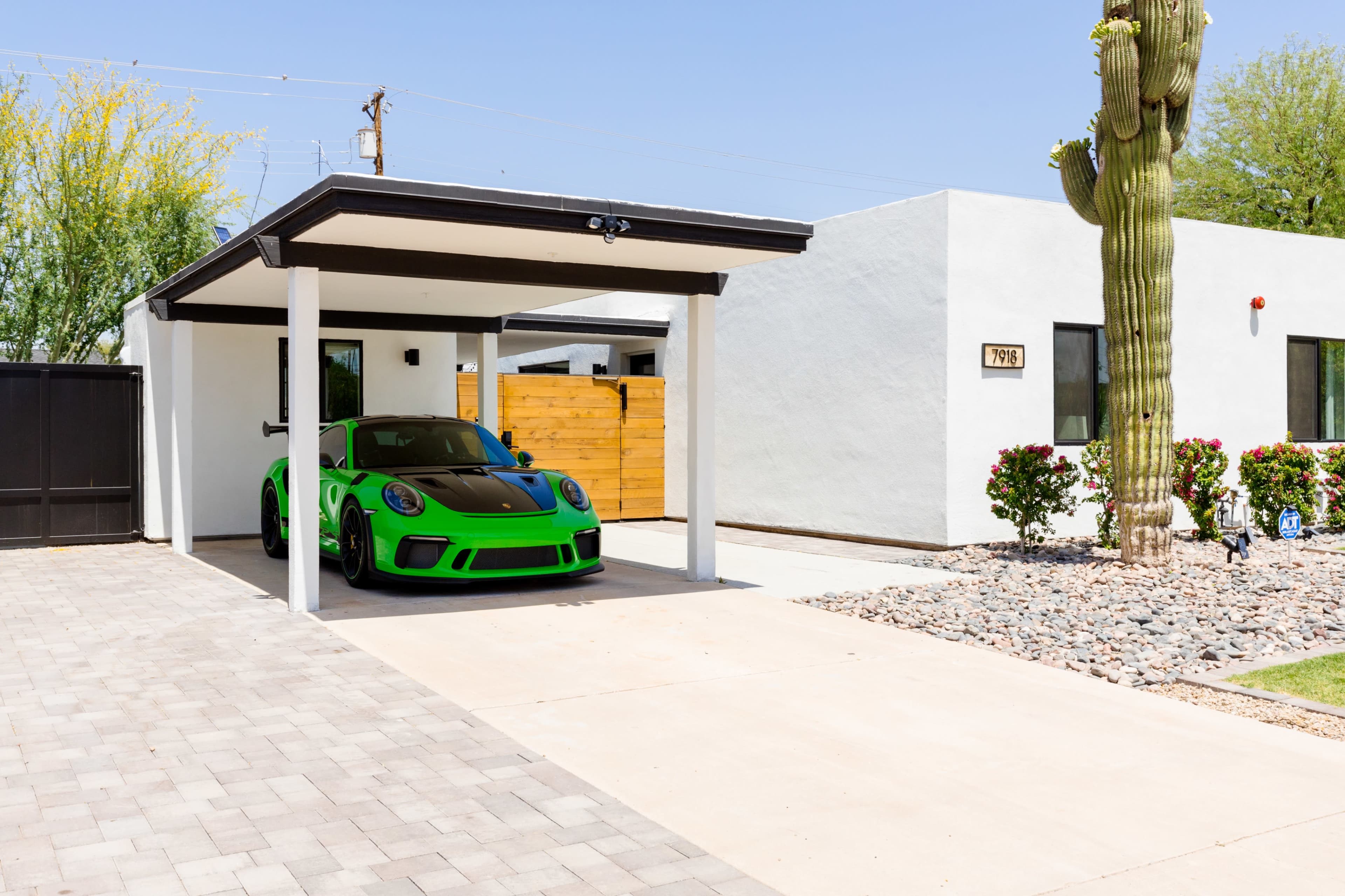 A bright green sports car is parked under a carport in front of a modern white house with a landscaped yard featuring a cactus and gravel.