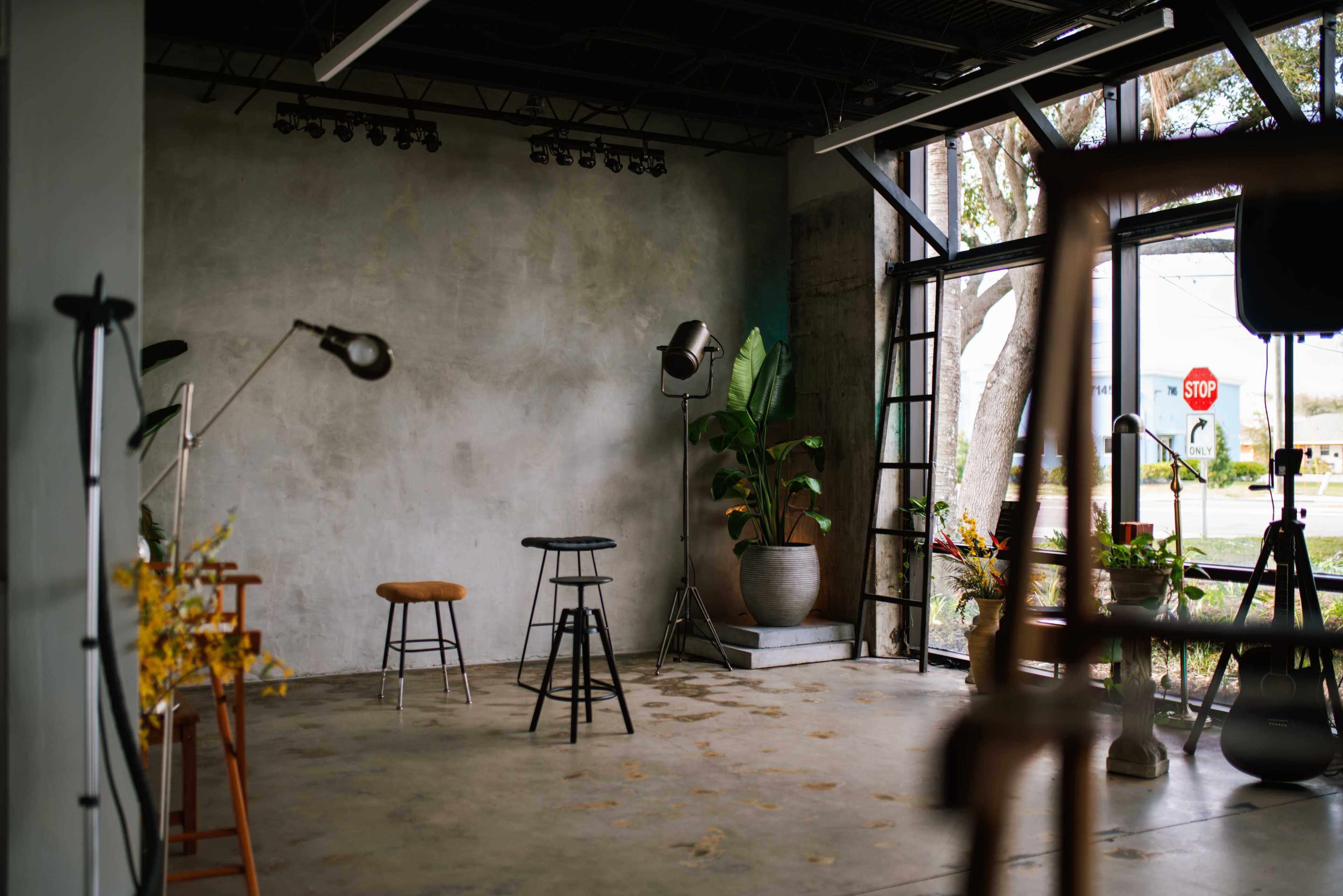 The image depicts a minimalist studio space with exposed concrete walls, several chairs, plants in pots, and adjustable lighting equipment.