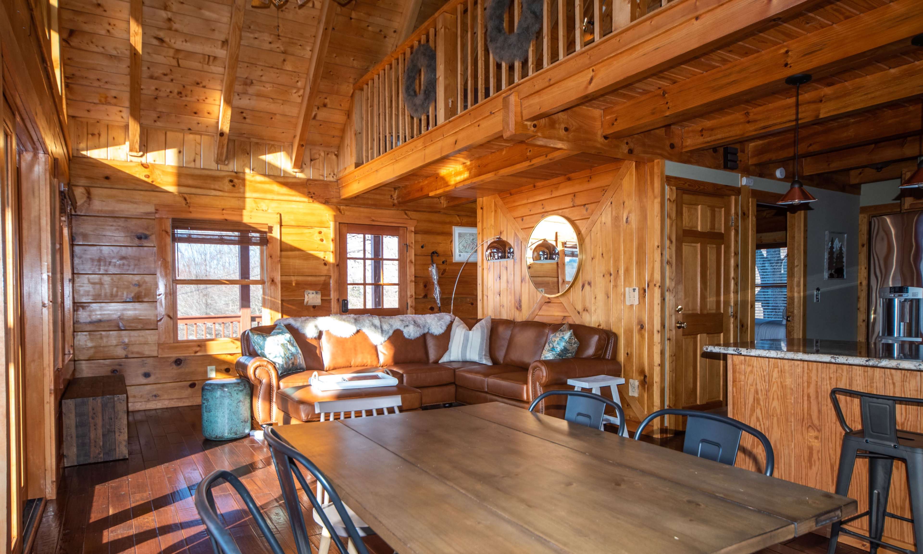 The interior of a wooden cabin features a leather sofa, a dining table, and large windows allowing sunlight to illuminate the space.