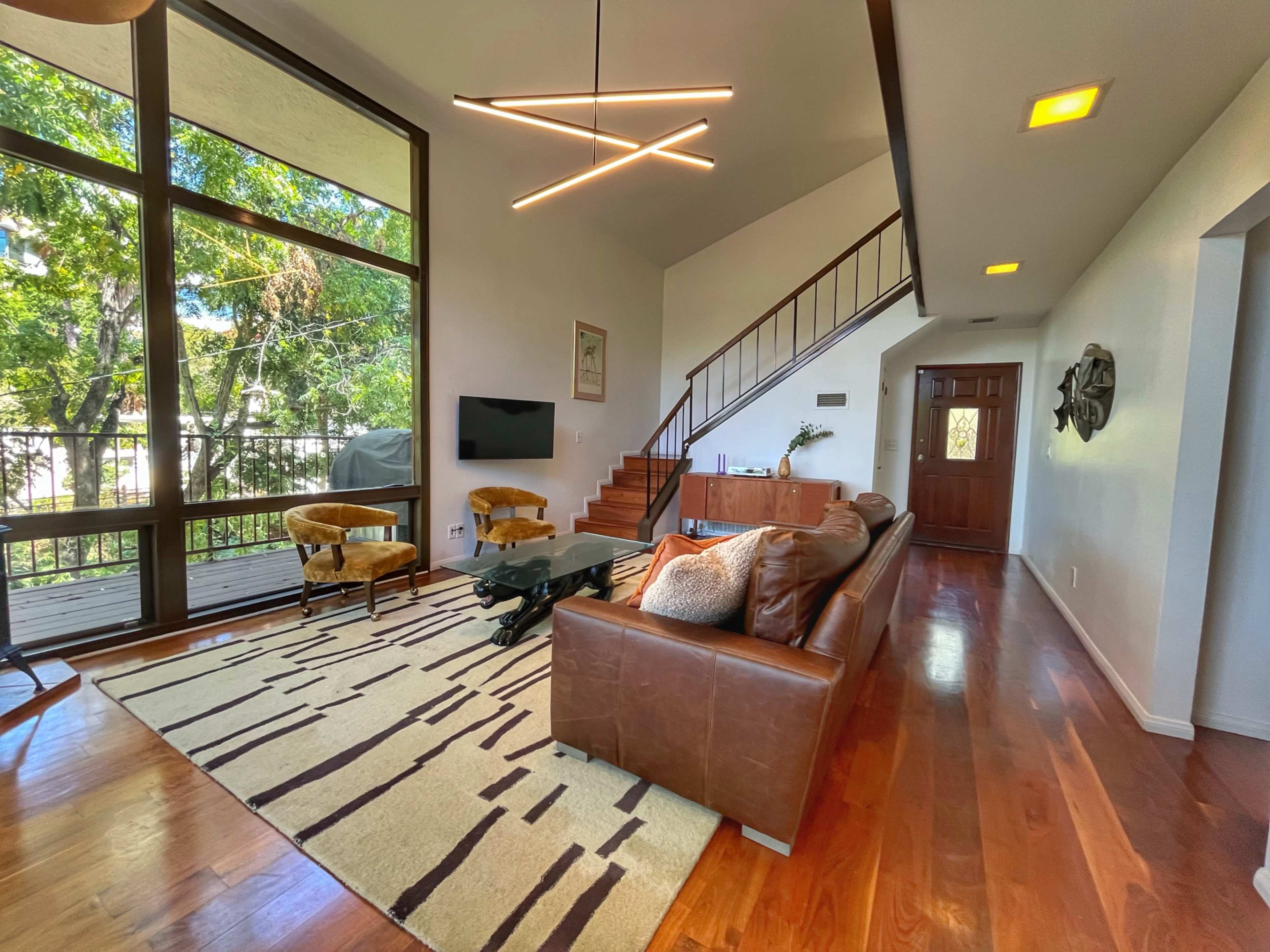 The image shows a modern living room with a brown leather sofa, a patterned rug, a glass coffee table, and large windows providing natural light.