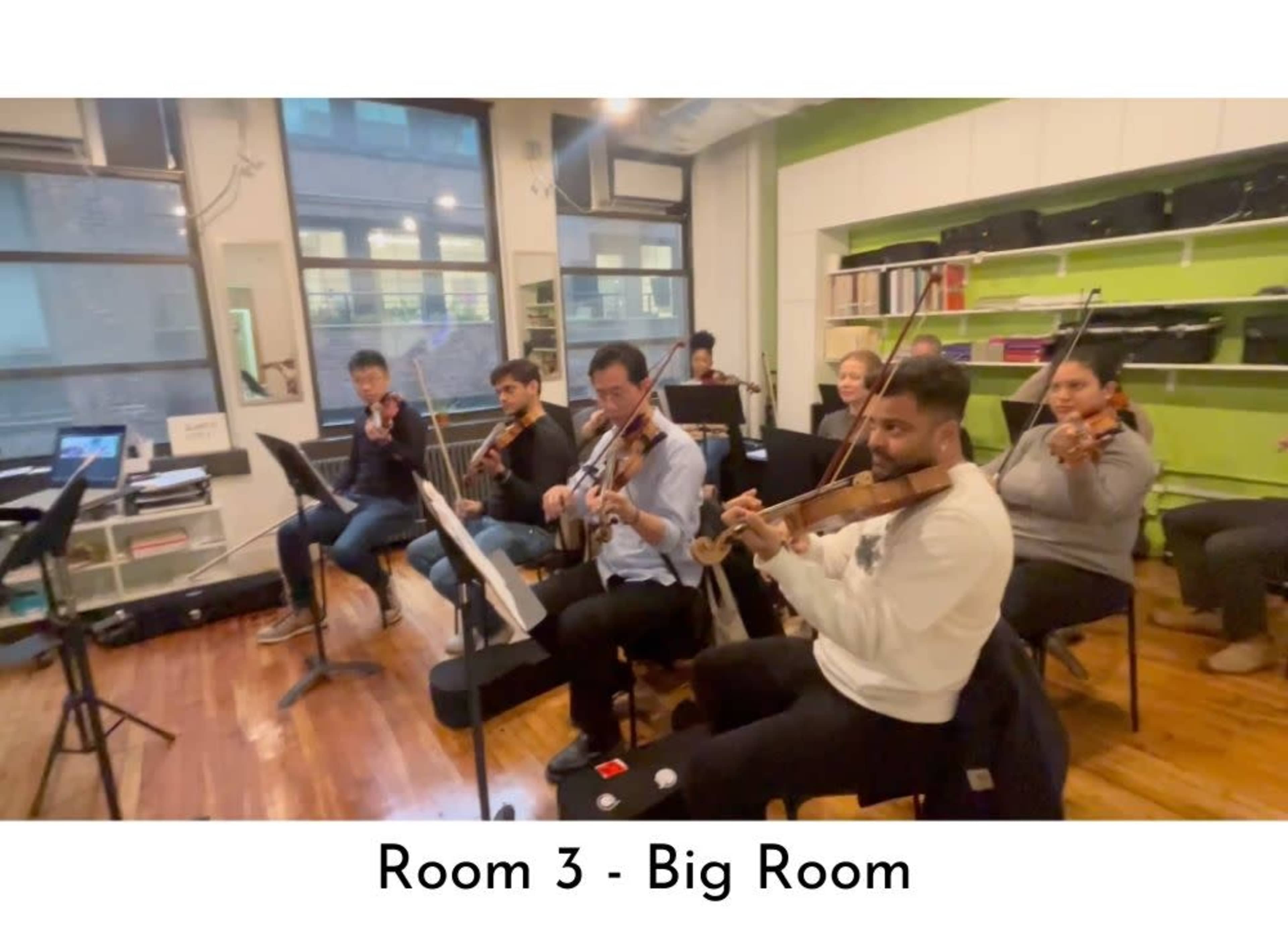 A group of musicians is seated in a large room, playing string instruments during a rehearsal session.