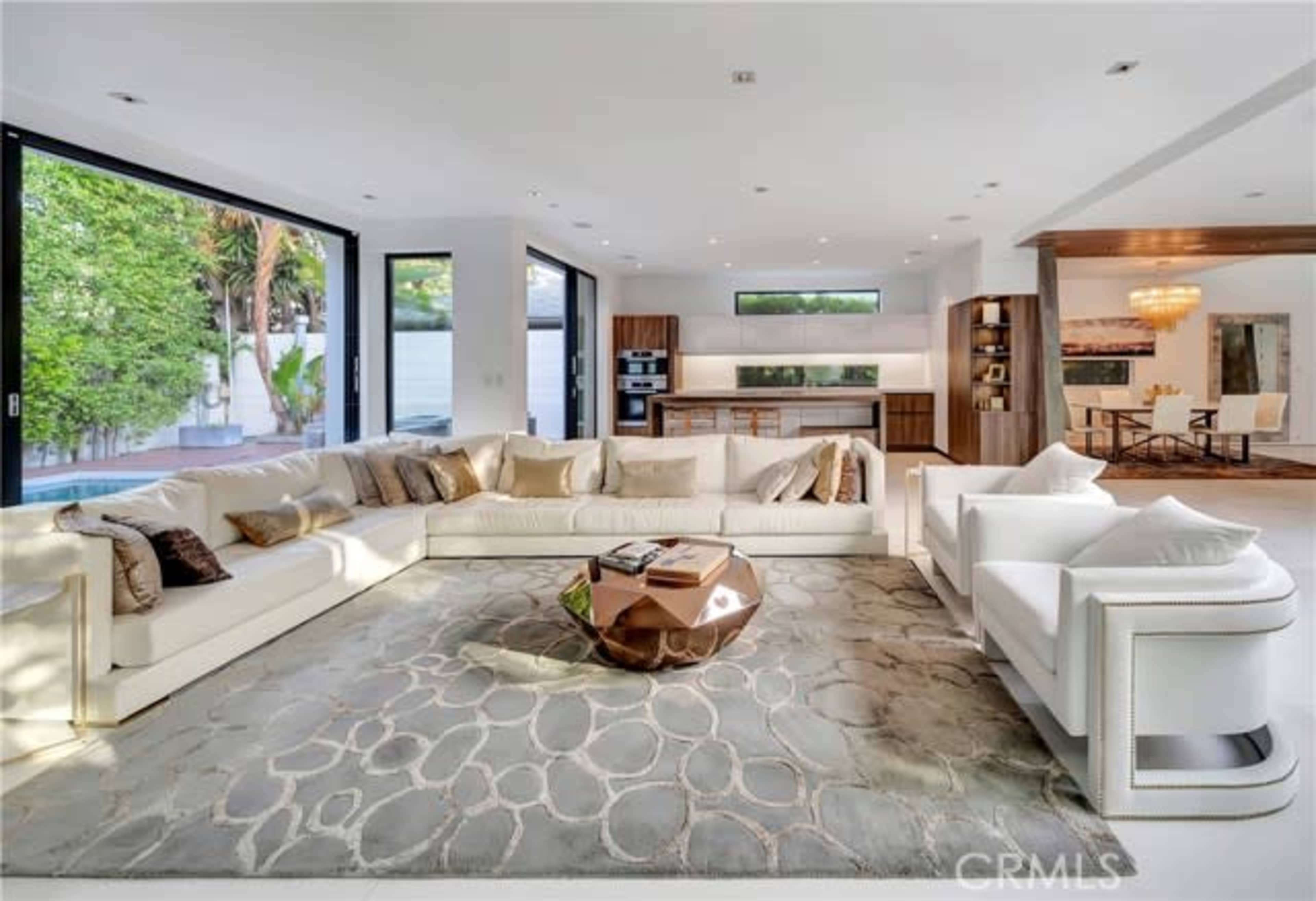 The living room features a large white sectional sofa, a decorative coffee table, and floor-to-ceiling windows overlooking a lush outdoor space.