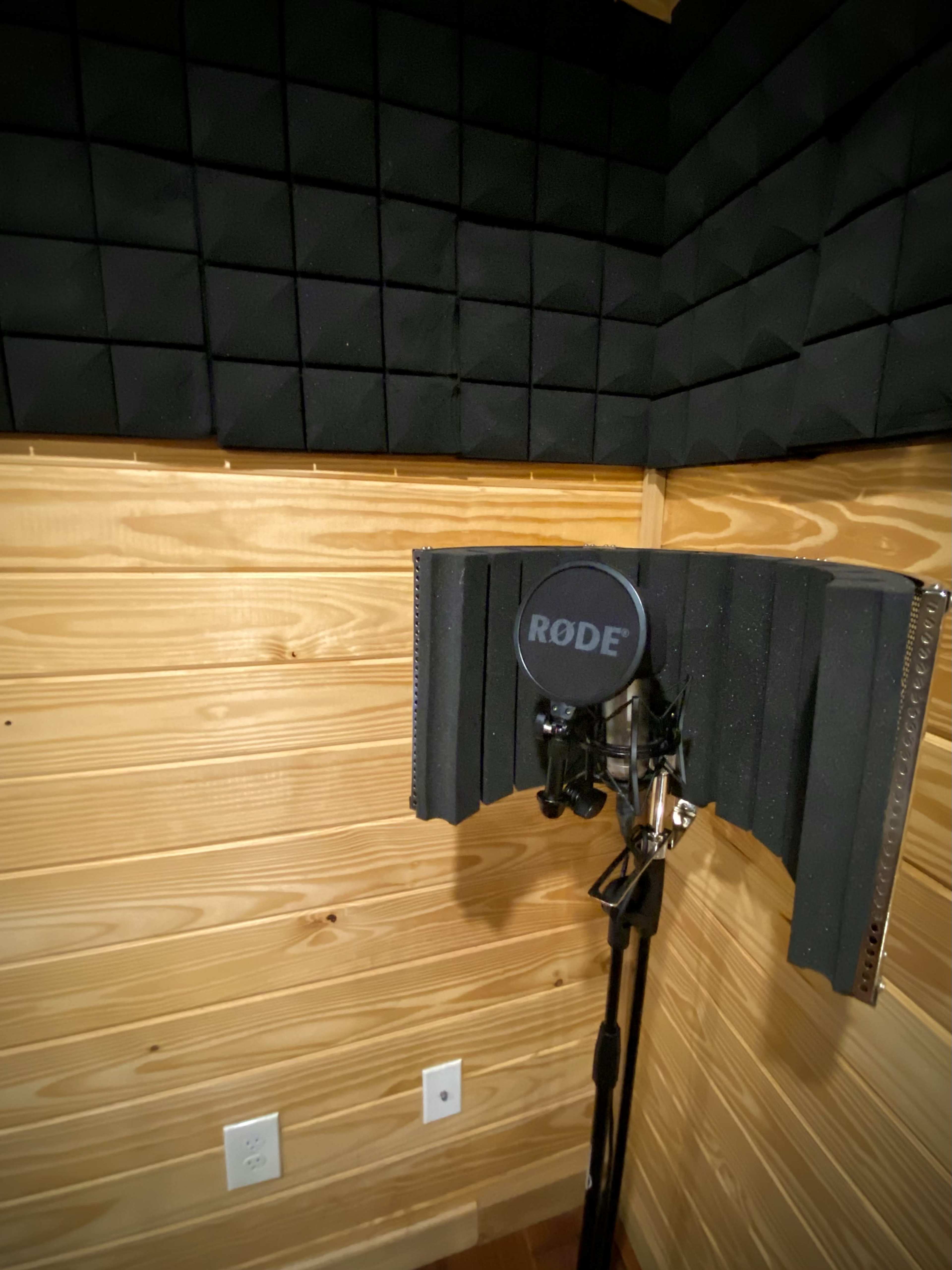 The image shows a microphone with a pop filter positioned in a soundproof corner of a wooden room, featuring acoustic foam panels on the walls.