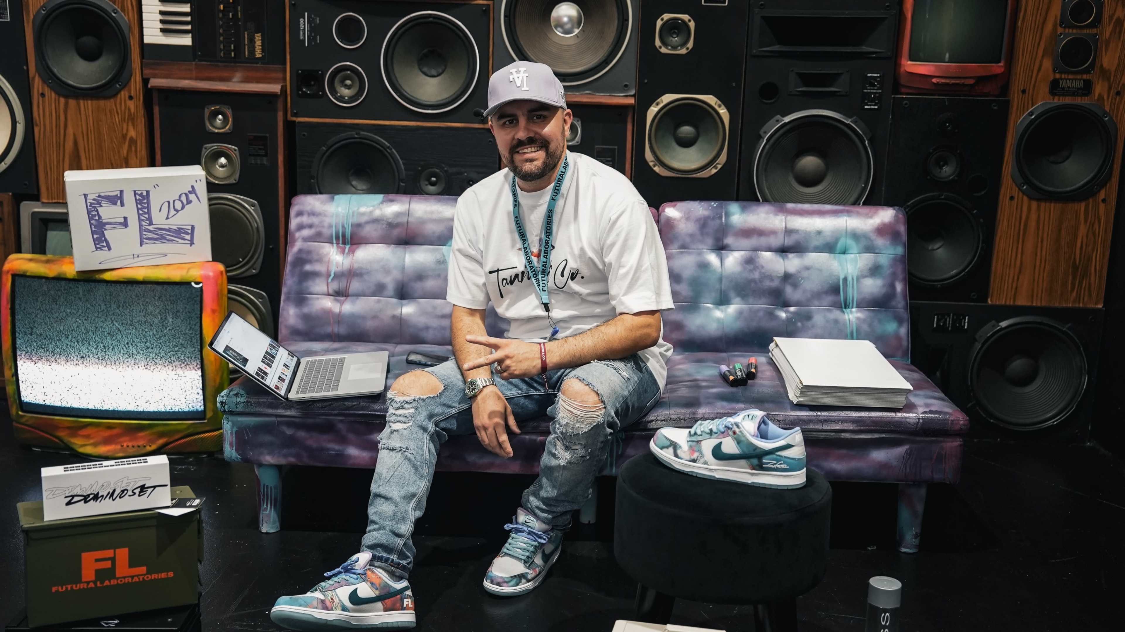 A man sits on a colorful couch in front of a wall of speakers, with a laptop and various items around him.