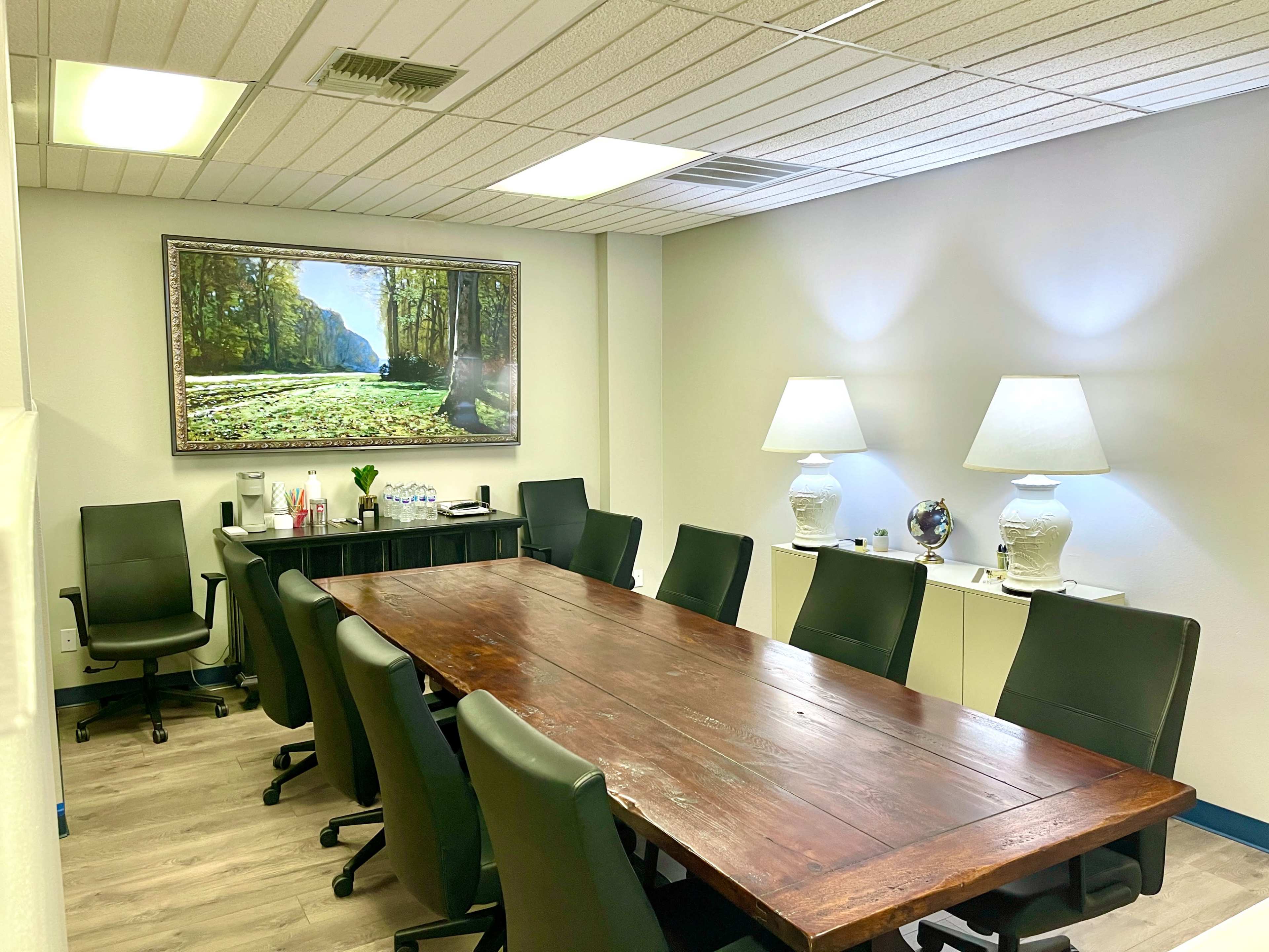 A conference room with a large wooden table, eight black chairs, two lamps, and a framed nature landscape painting on the wall.
