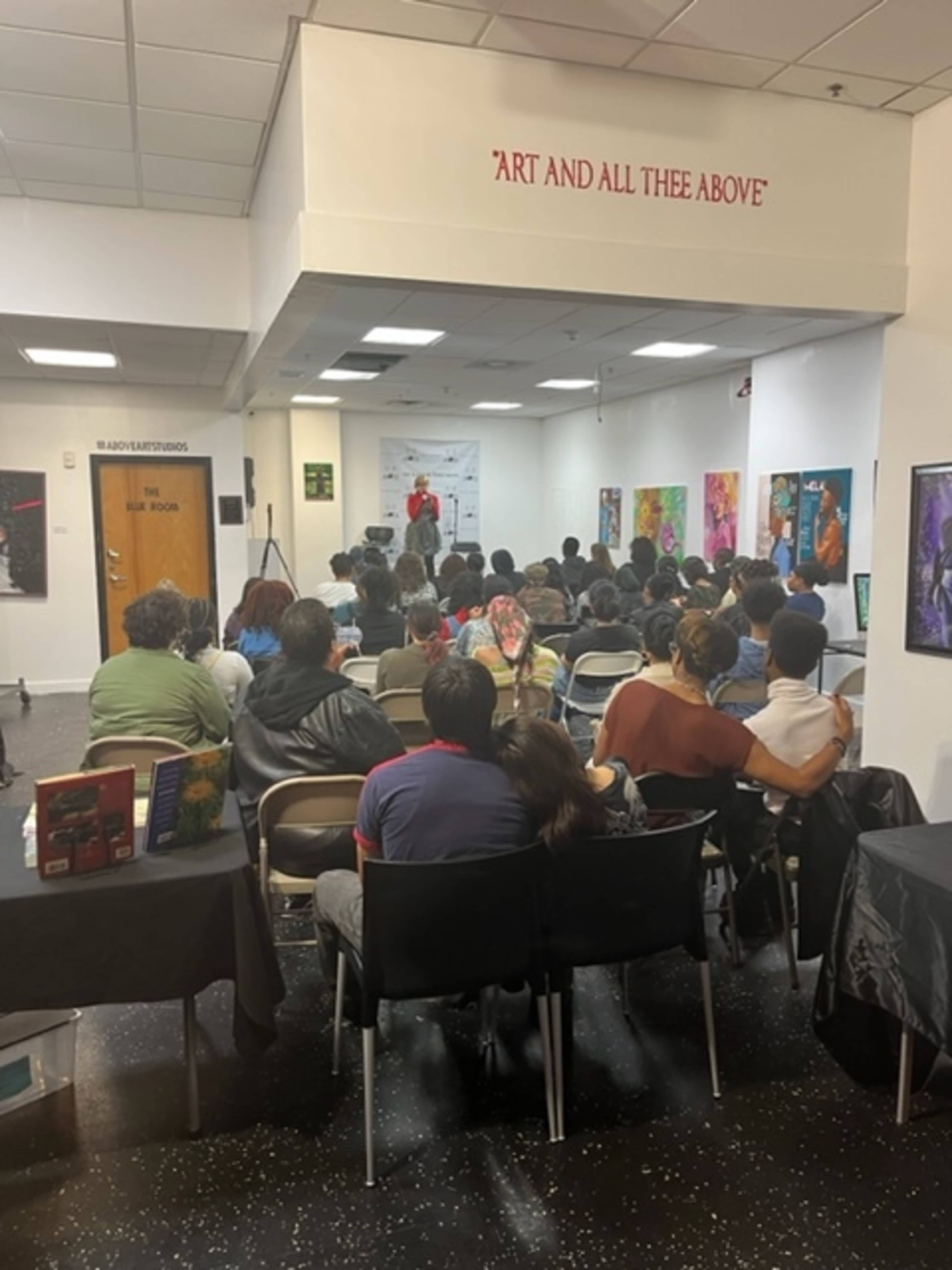 A speaker addresses an audience seated in a gallery with artwork on the walls.