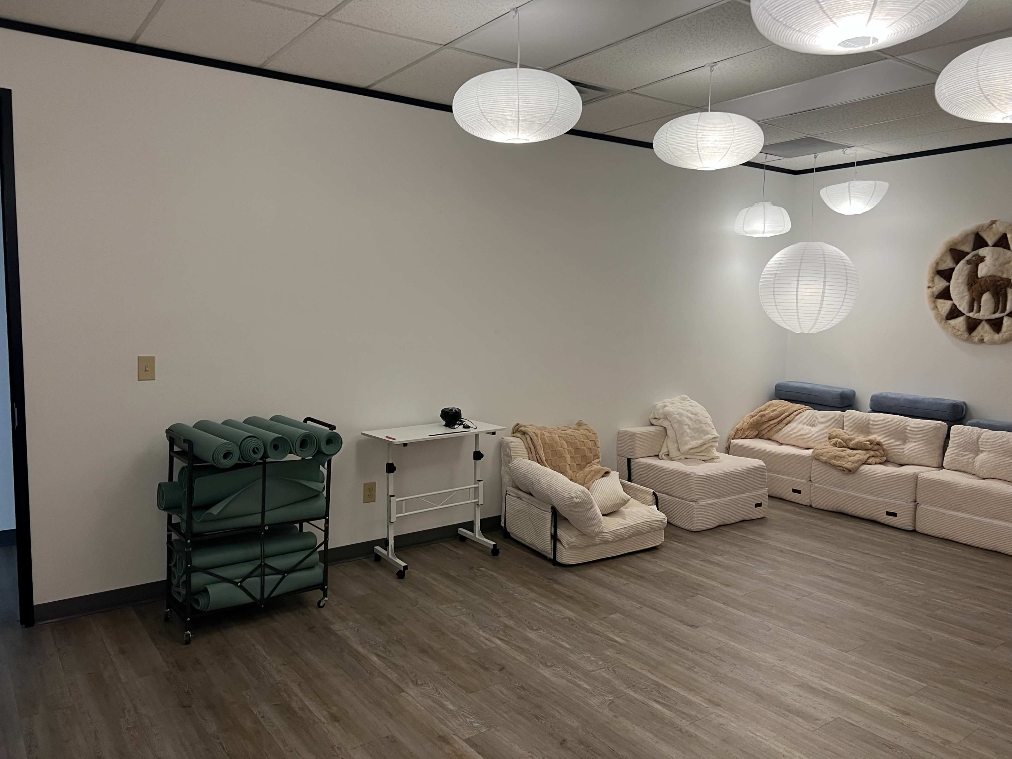 The image shows a spacious, minimalist room with light-colored walls, several hanging paper lanterns, a stack of yoga mats on a cart, and soft seating arranged along one wall.