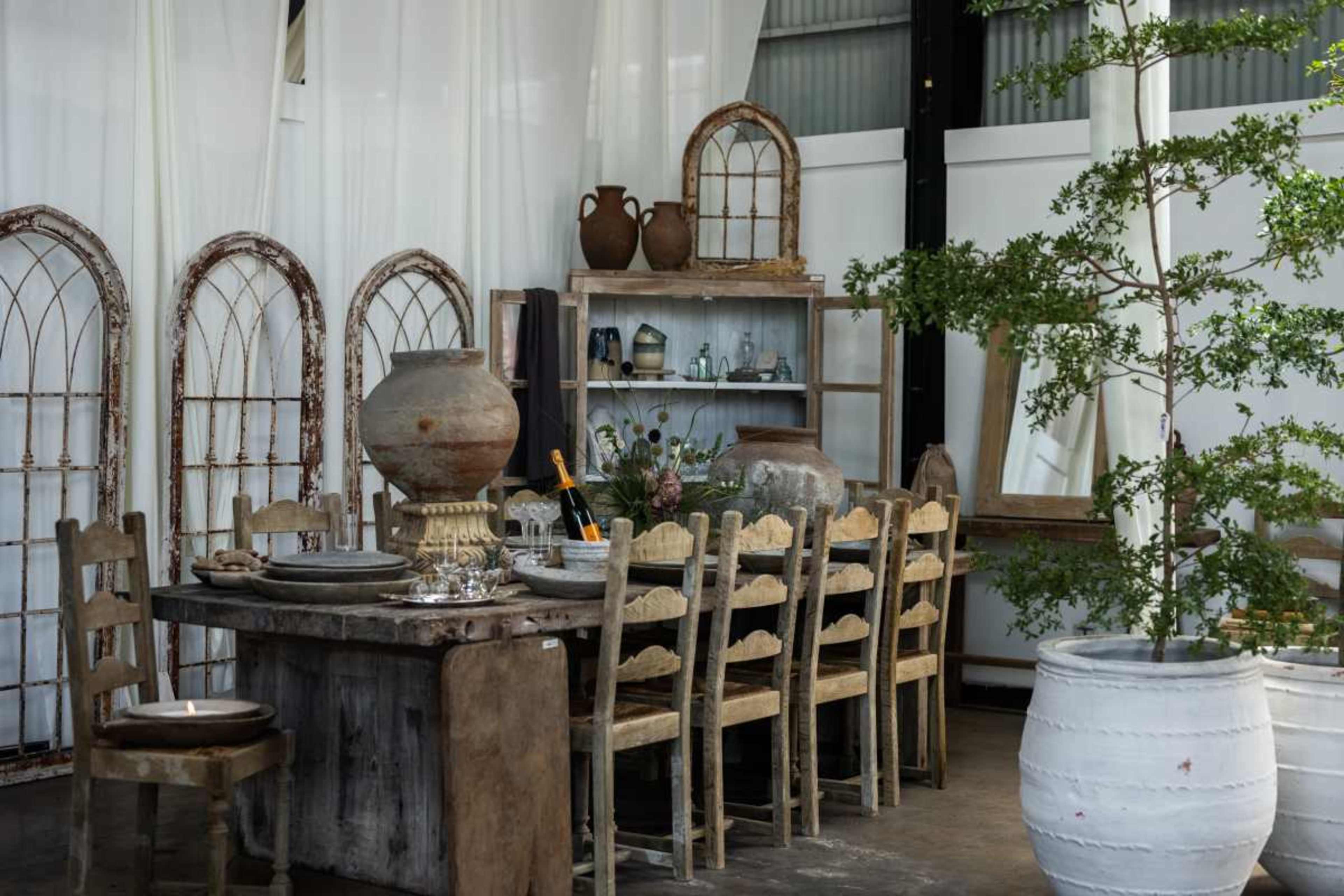 A rustic dining area features a large wooden table surrounded by chairs, with a variety of pottery and decor elements on display, including potted greenery and decorative mirrors.