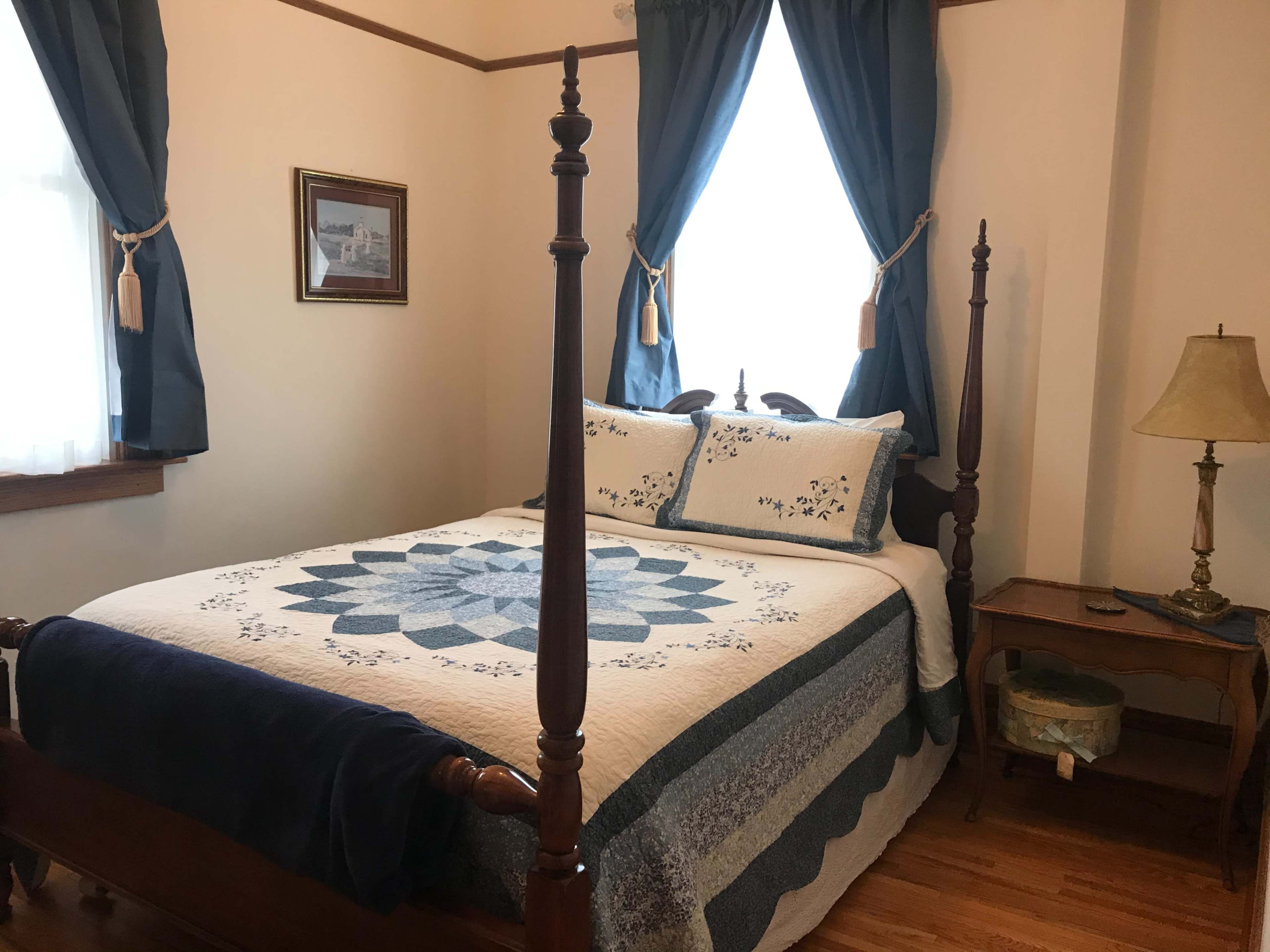 A bedroom with a four-poster bed covered by a quilt, a bedside table with a lamp, and blue curtains framing a window.
