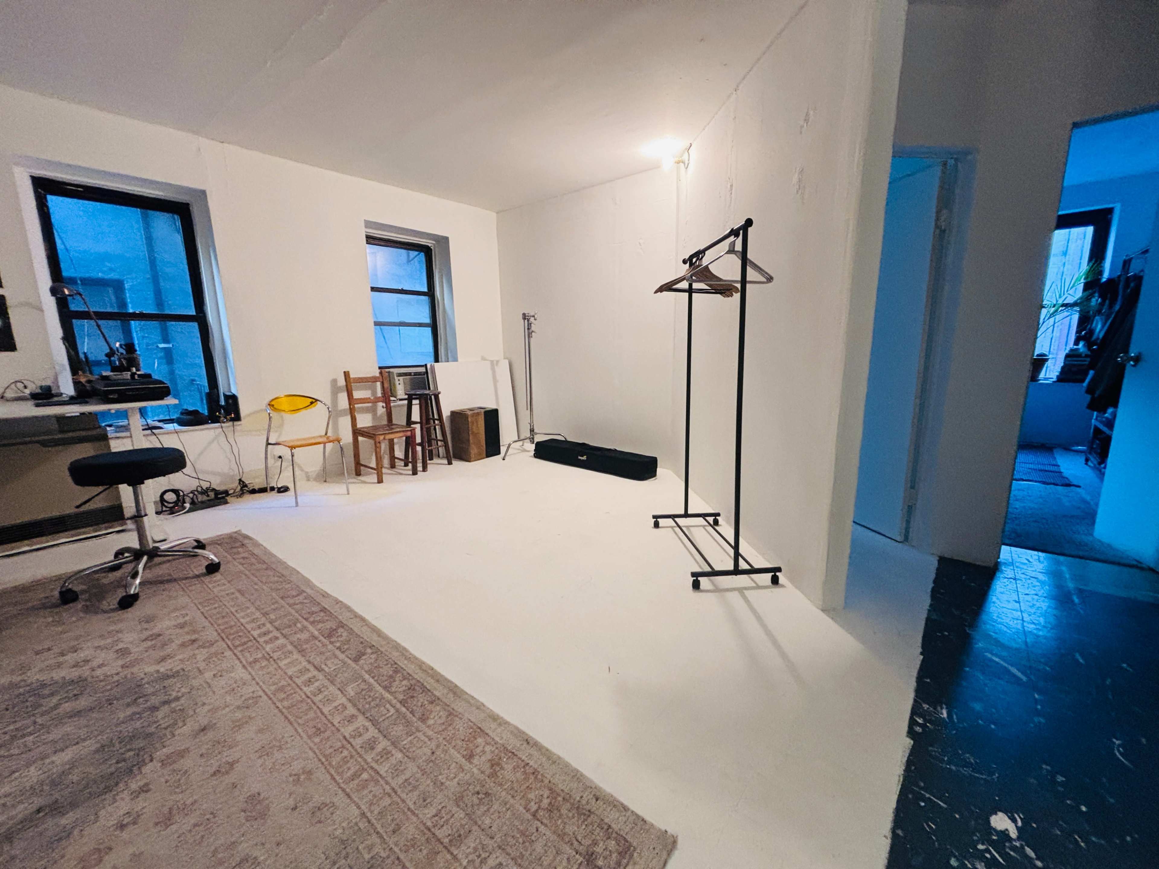The image shows a minimalistic room with white walls, a wooden rug, a desk with a chair, a clothes rack, and a doorway leading to another room.