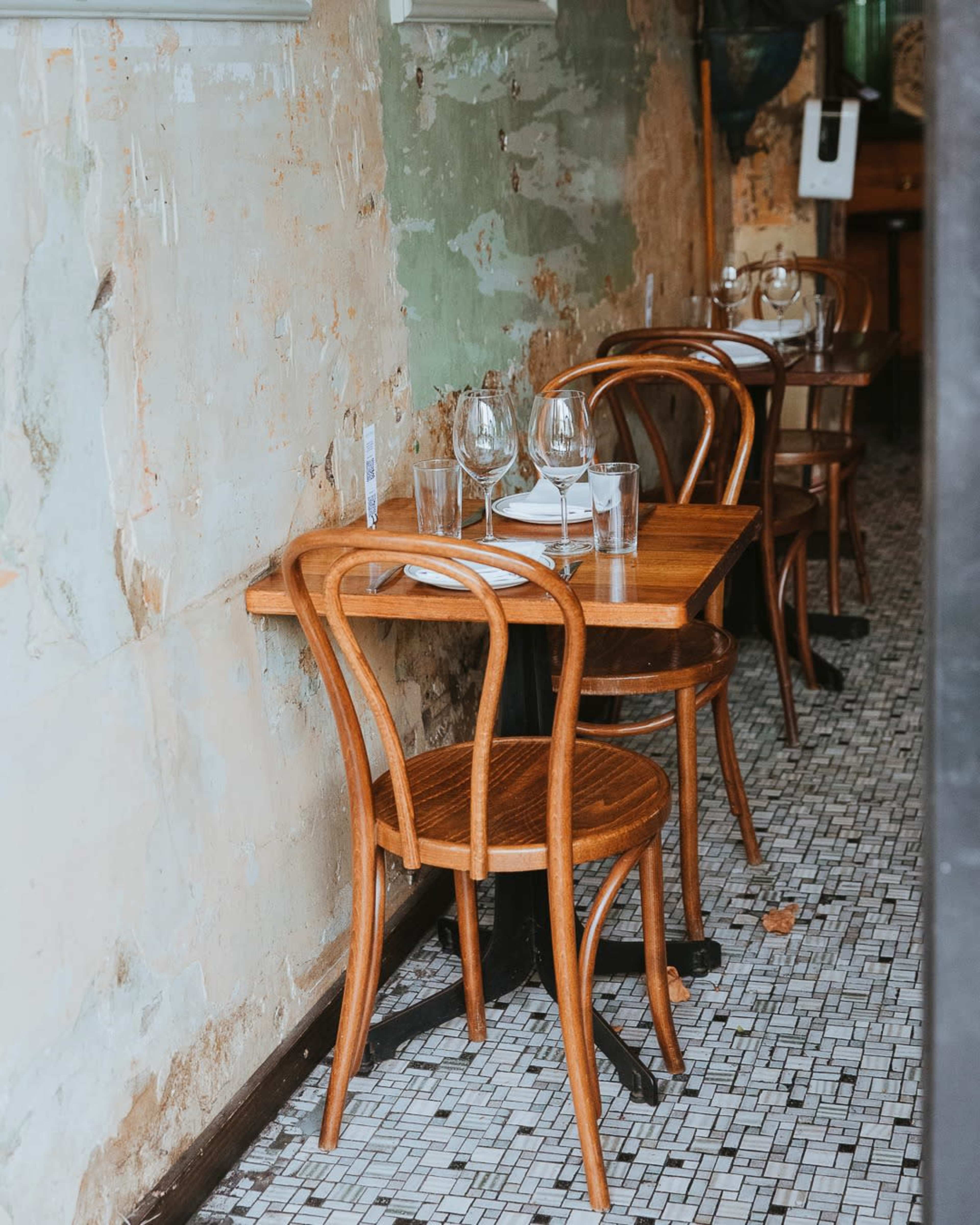 A small wooden table is set with glasses and plates in a restaurant featuring exposed walls and tiled flooring.