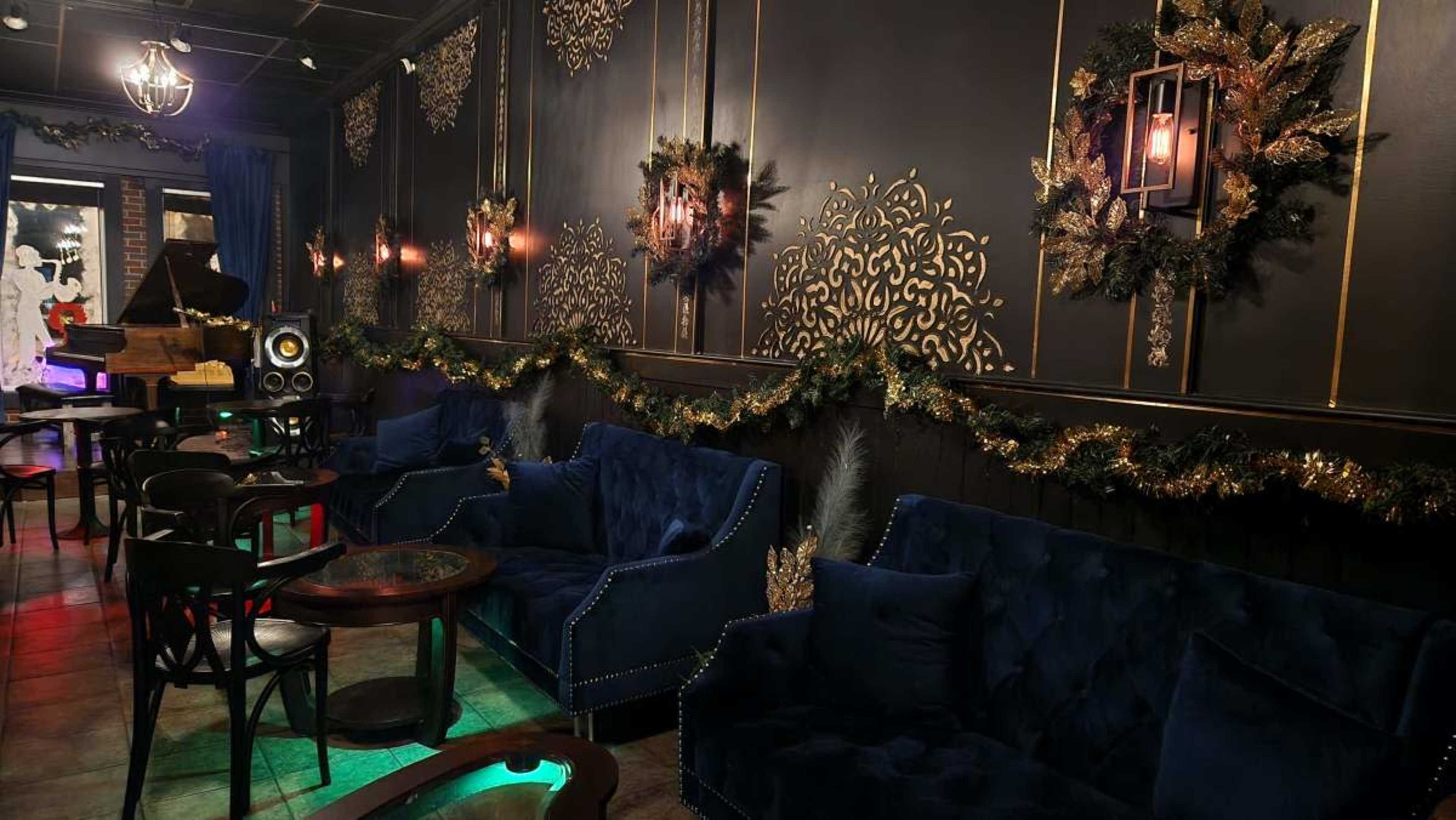 The image shows a dimly lit lounge with dark walls adorned with decorative gold patterns, festive wreaths, and plush blue seating arranged around small tables.