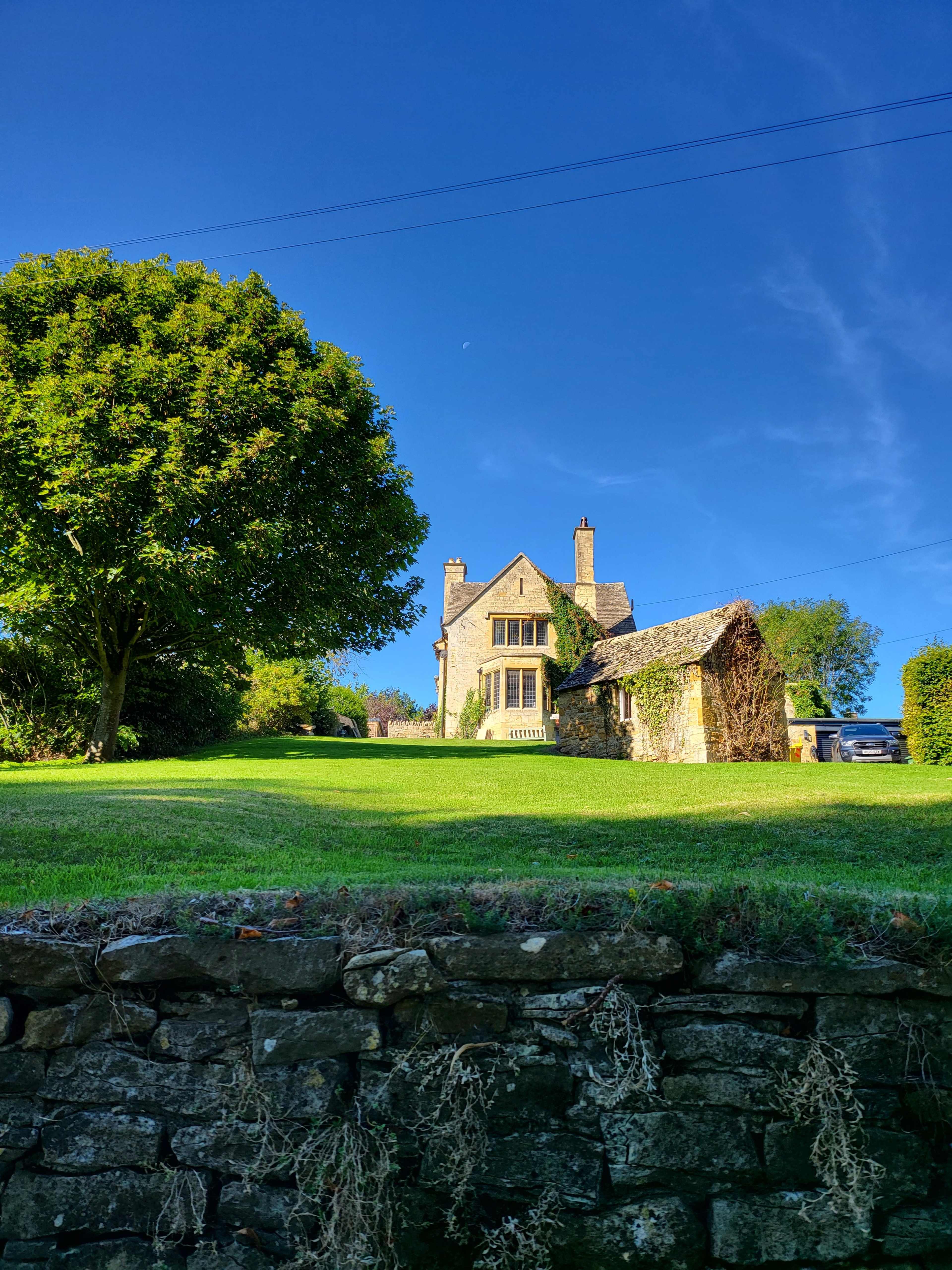 A stone house with tall windows sits on a green hillside, surrounded by trees under a clear blue sky.
