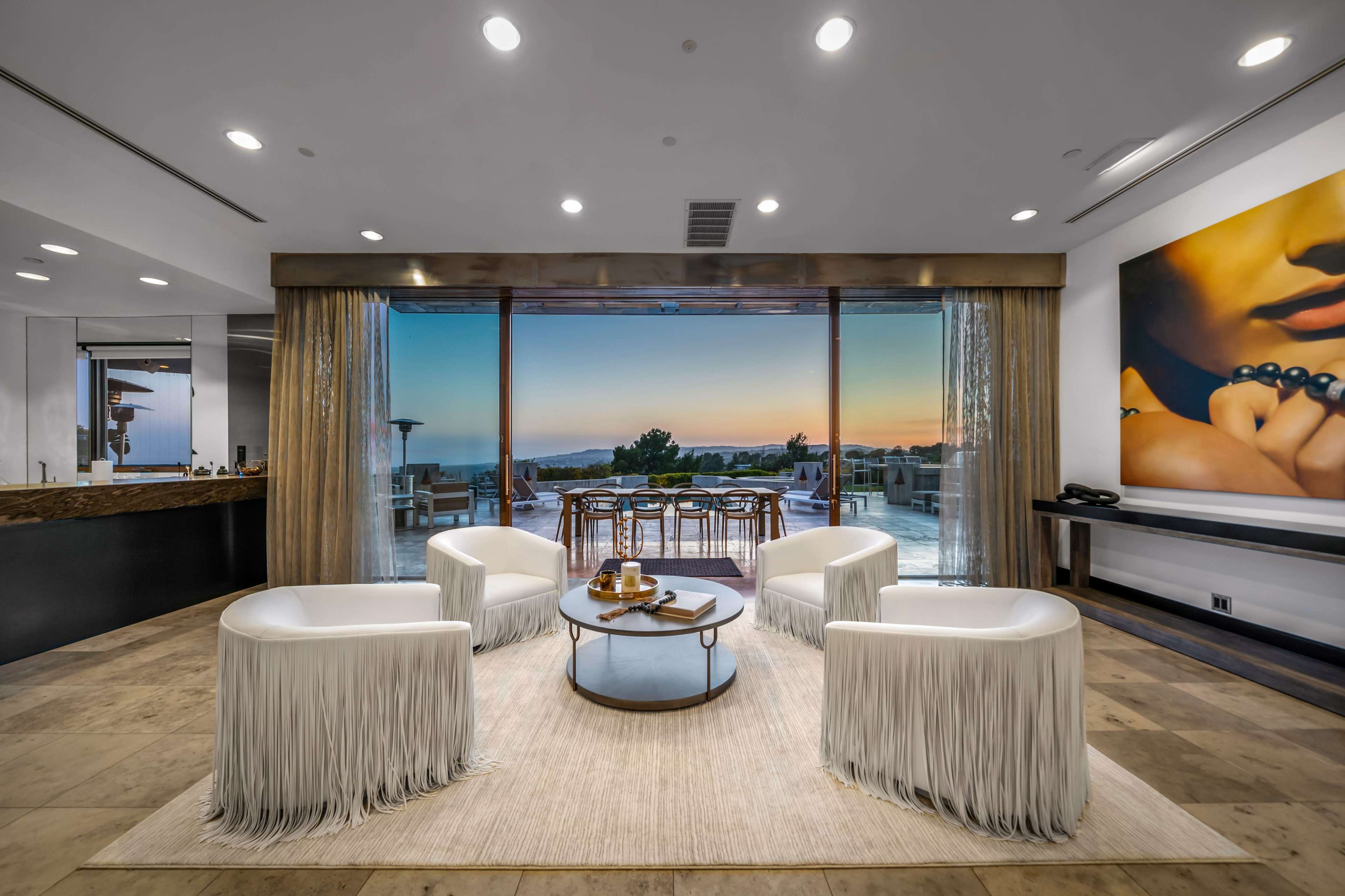 A modern living space features four white chairs with fringe, a round coffee table, and large windows showing a sunset view over a balcony.