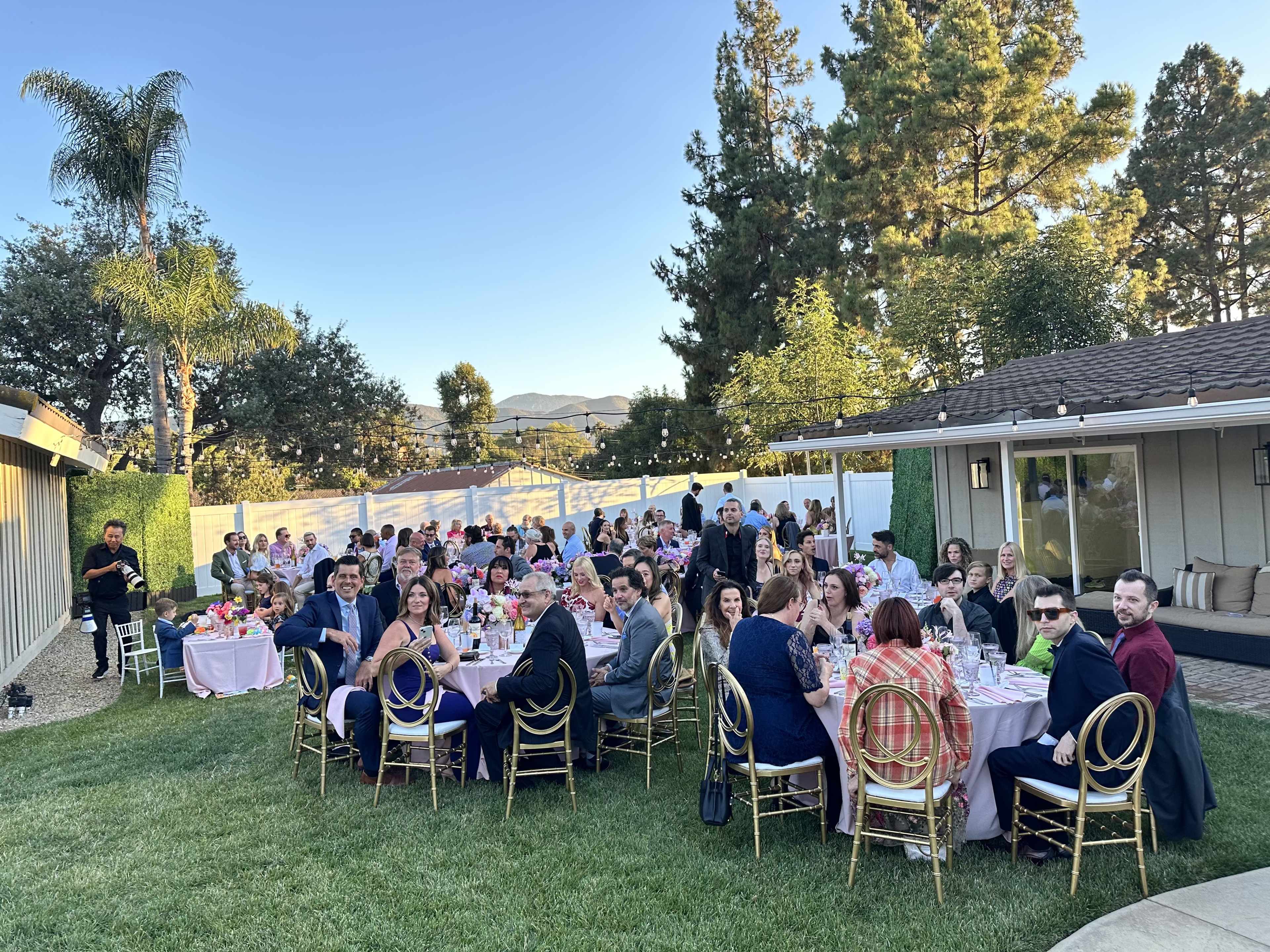 A large group of people is seated at elegantly set tables in a grassy outdoor space, surrounded by trees and a residential building in the background.