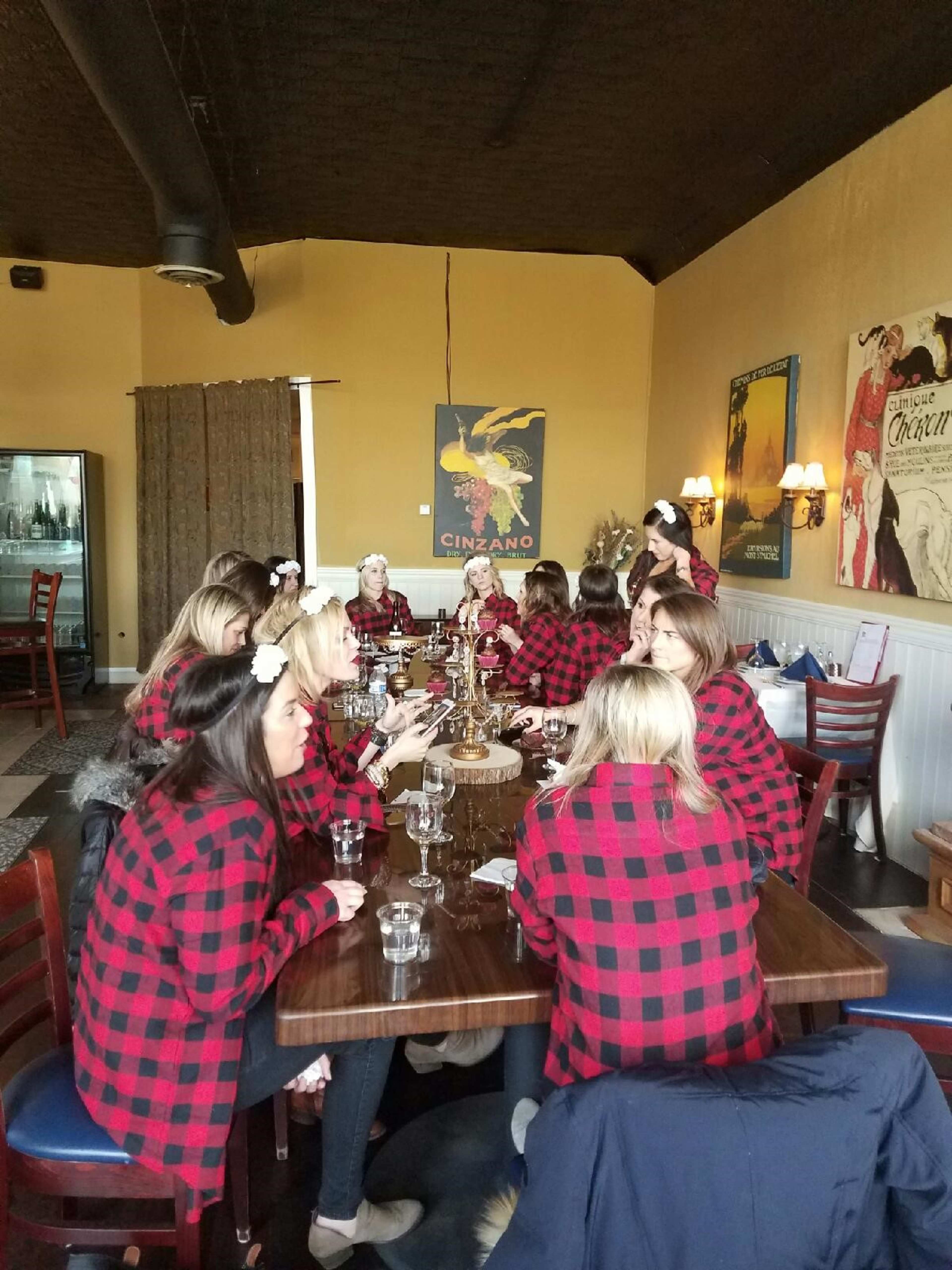 A group of women wearing matching red and black plaid flannel shirts sit around a large wooden table in a restaurant.
