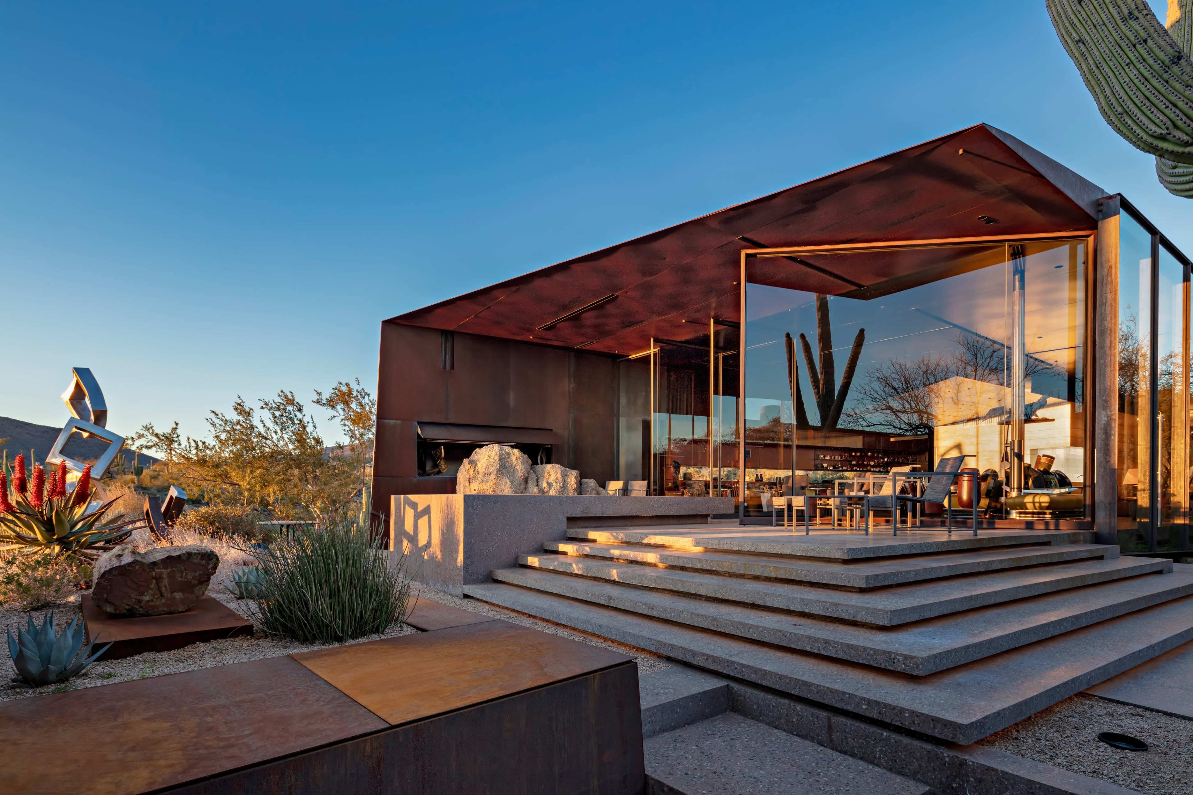 A modern house with a metal exterior and large glass windows is situated in a desert landscape, featuring an outdoor seating area and surrounding cacti.
