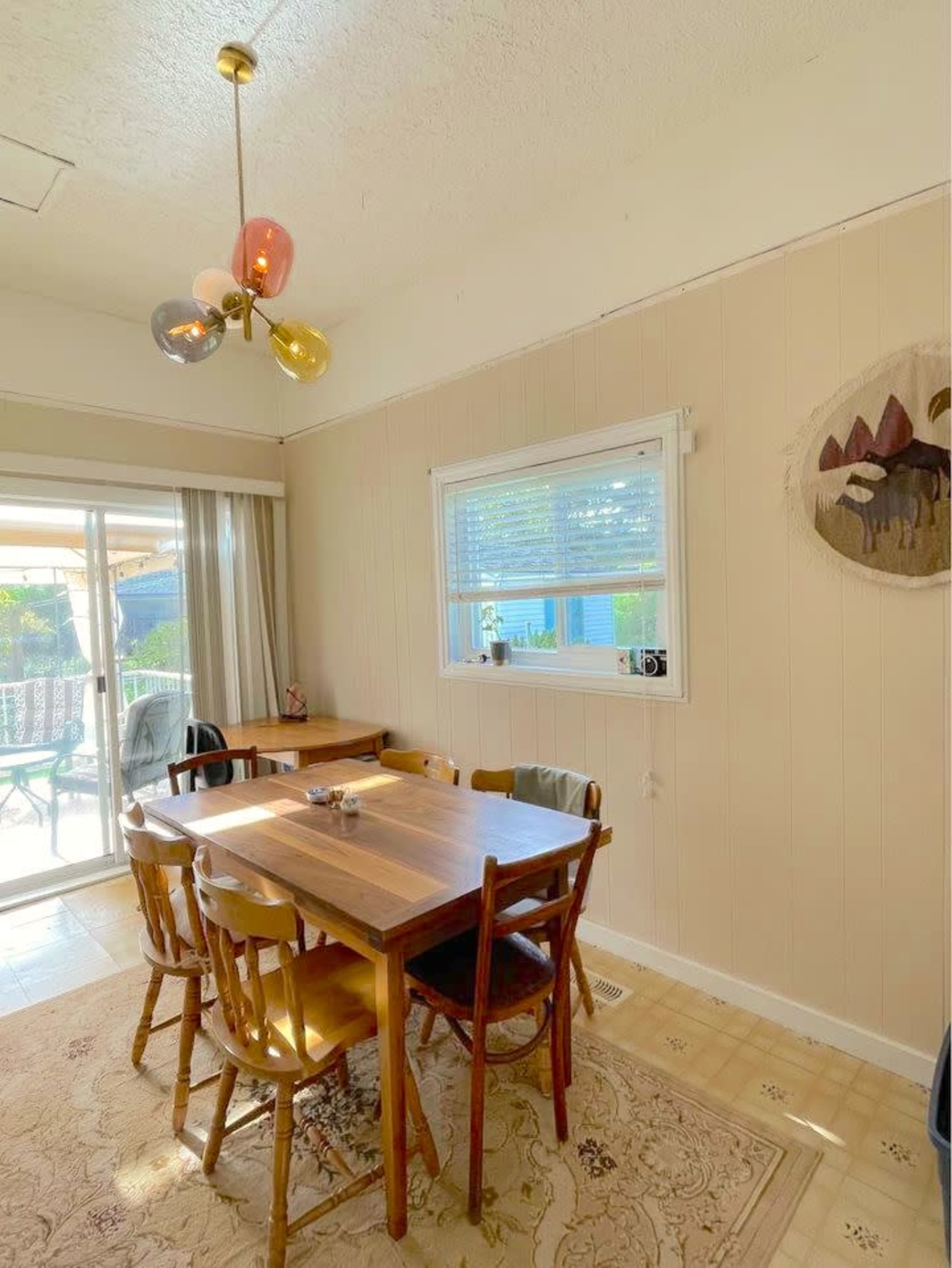 A dining area features a wooden table surrounded by chairs, with a colorful light fixture above and a window revealing an outdoor view.