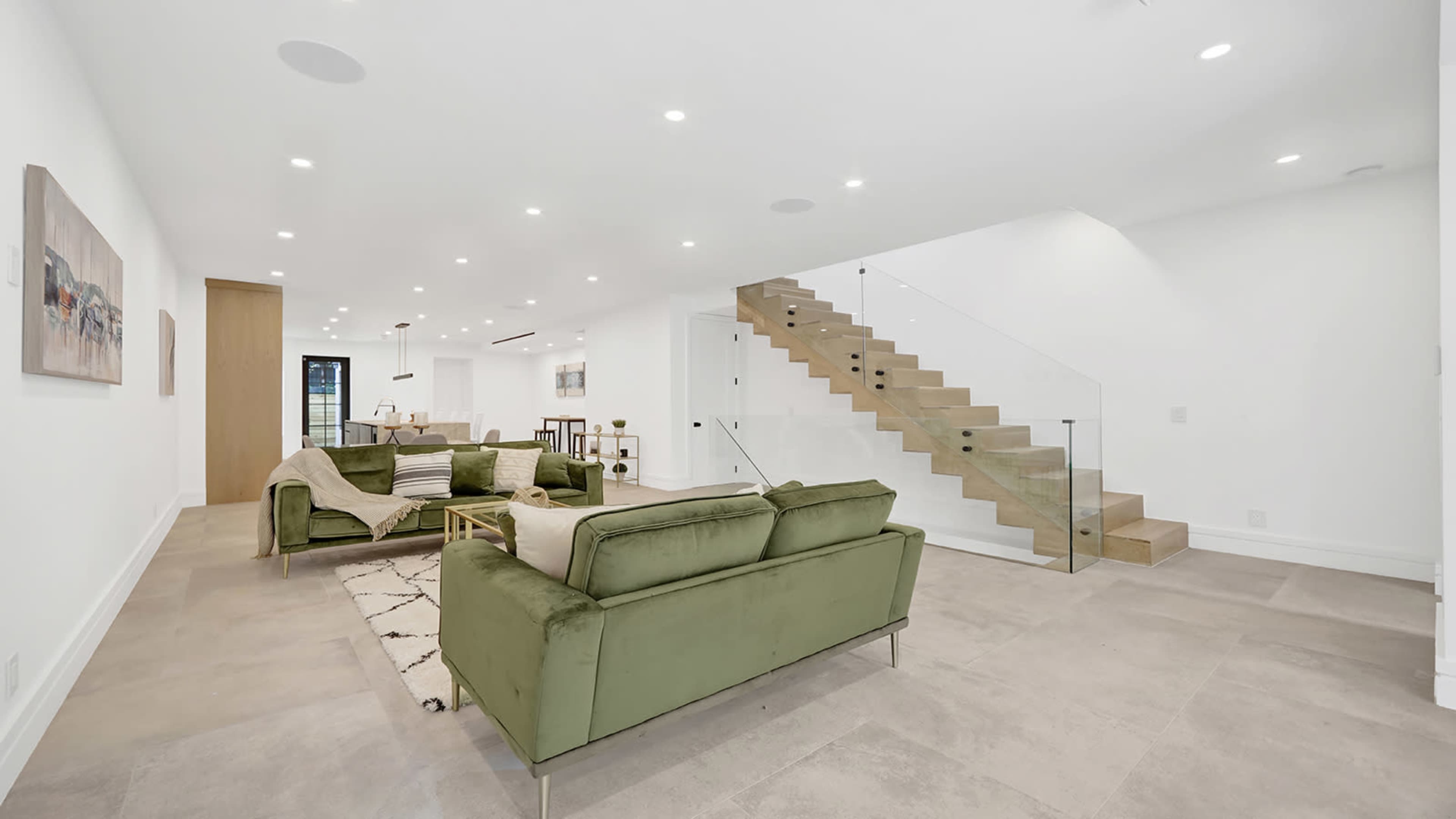 A modern living room with green sofas, a patterned rug, and a staircase with glass railings leading to an upper level.