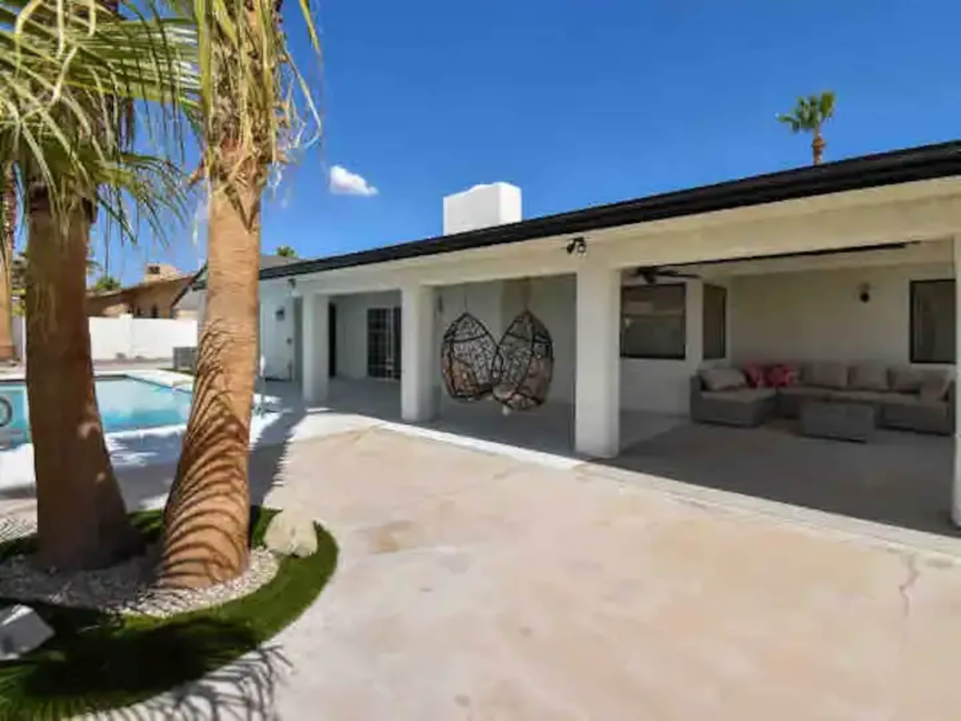 A modern backyard with a swimming pool, palm trees, and a covered seating area featuring a hanging chair.