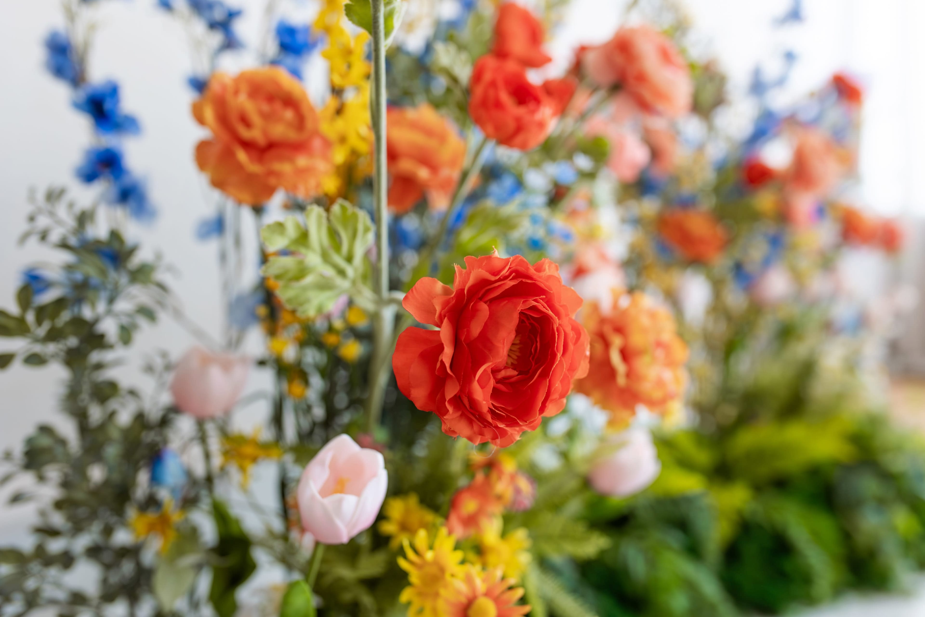 The image shows a vibrant arrangement of various flowers, including orange roses, pink tulips, and blue delphiniums, set against a blurred background of greenery.