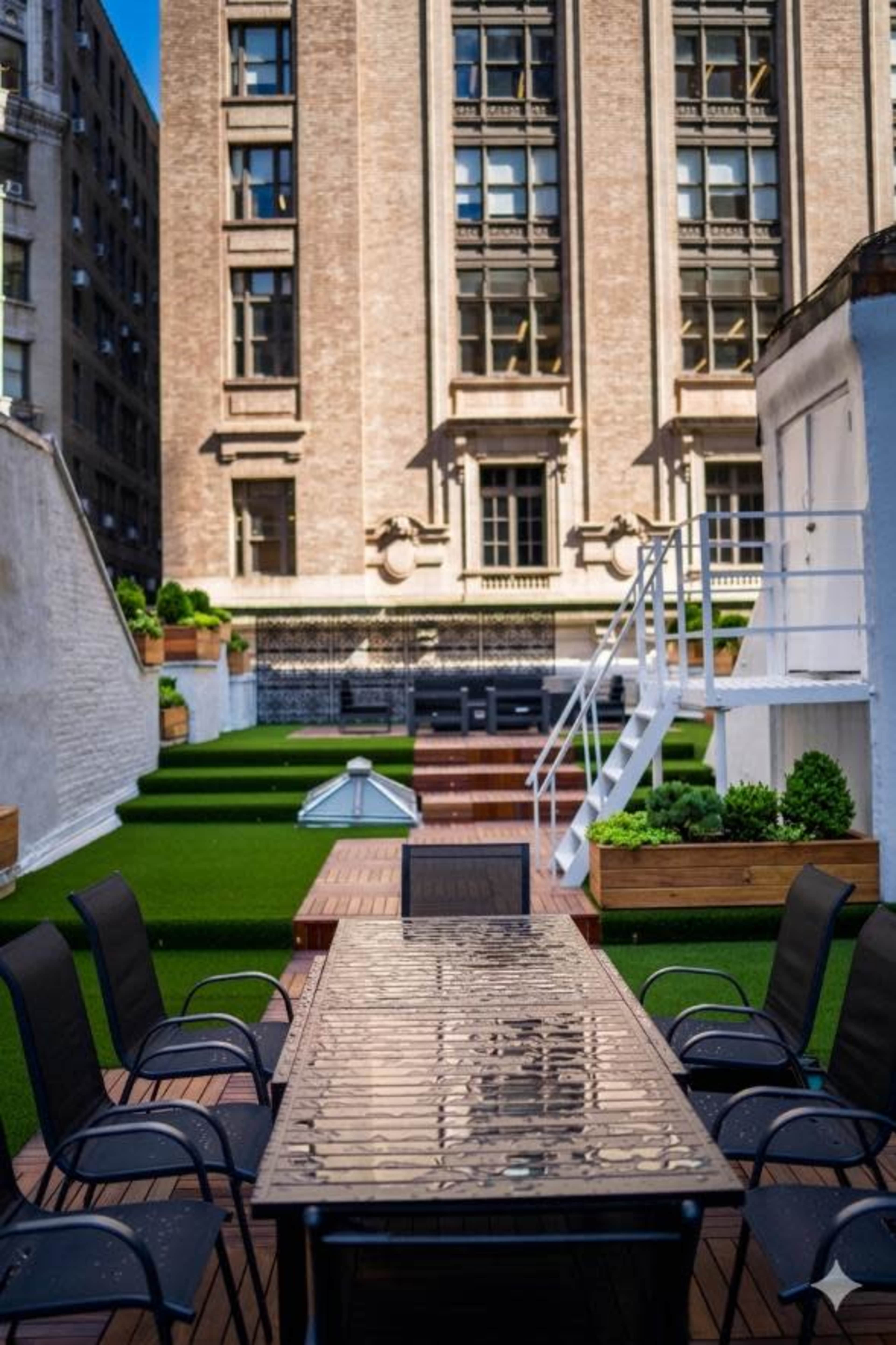 The image shows a rooftop terrace with a long table surrounded by black chairs, grass-like flooring, and planters, set against a backdrop of a brick building.