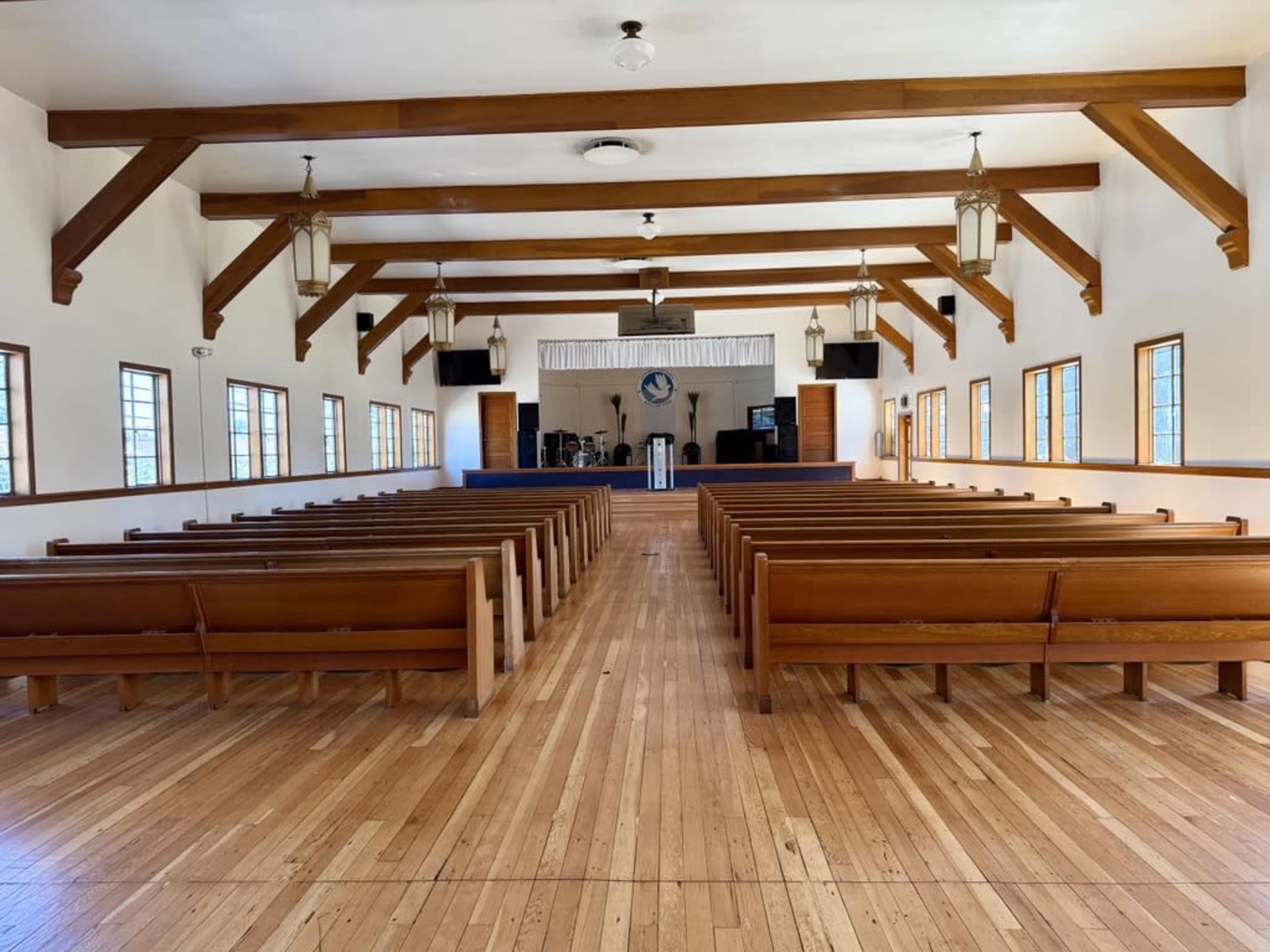 The interior of a church features rows of wooden pews facing a stage area at the front, with tall windows allowing natural light to enter.