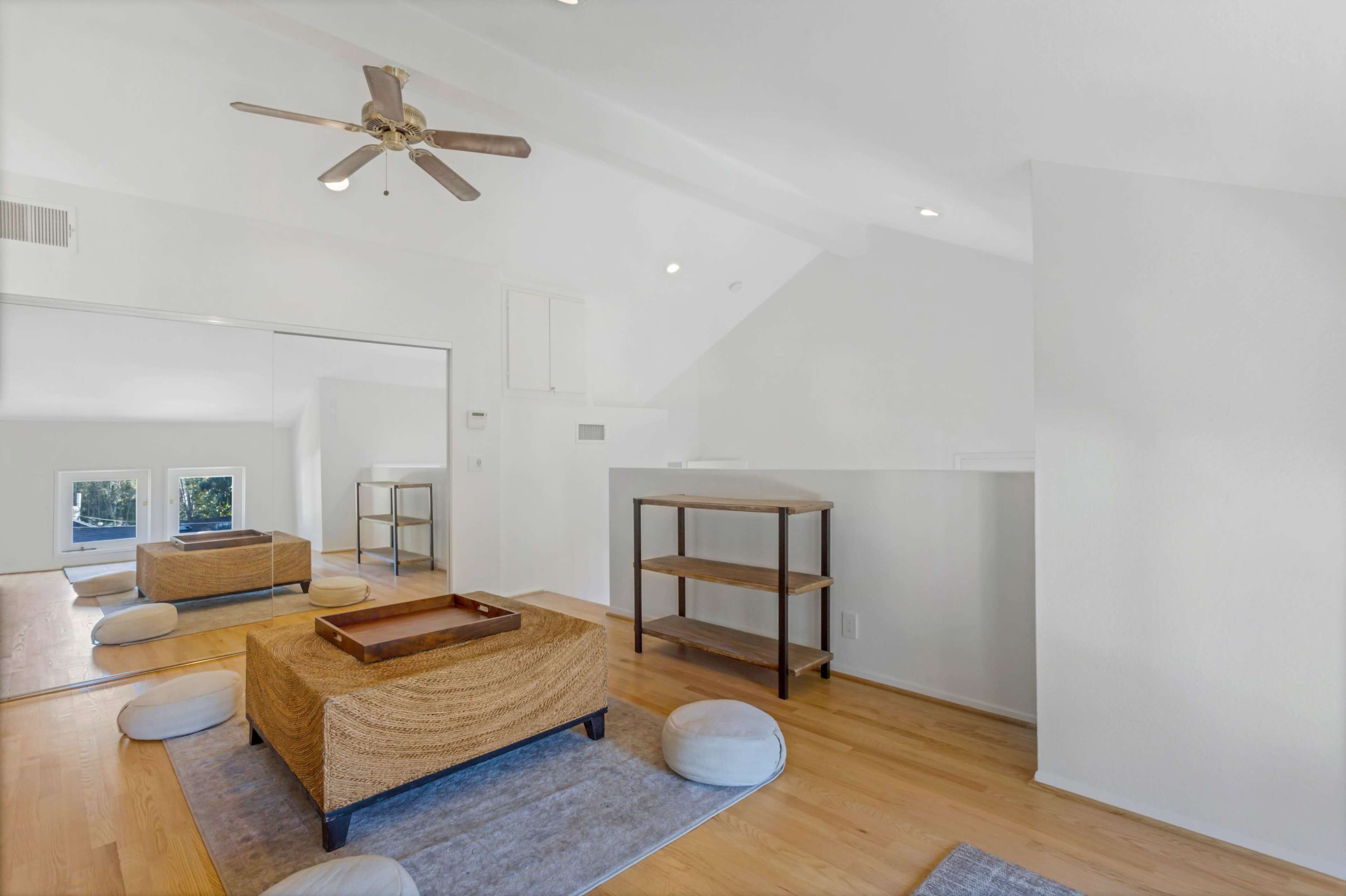 The image shows a bright, minimalist living space with a wooden floor, a low table, a shelving unit, and several cushions arranged on a light-colored rug.