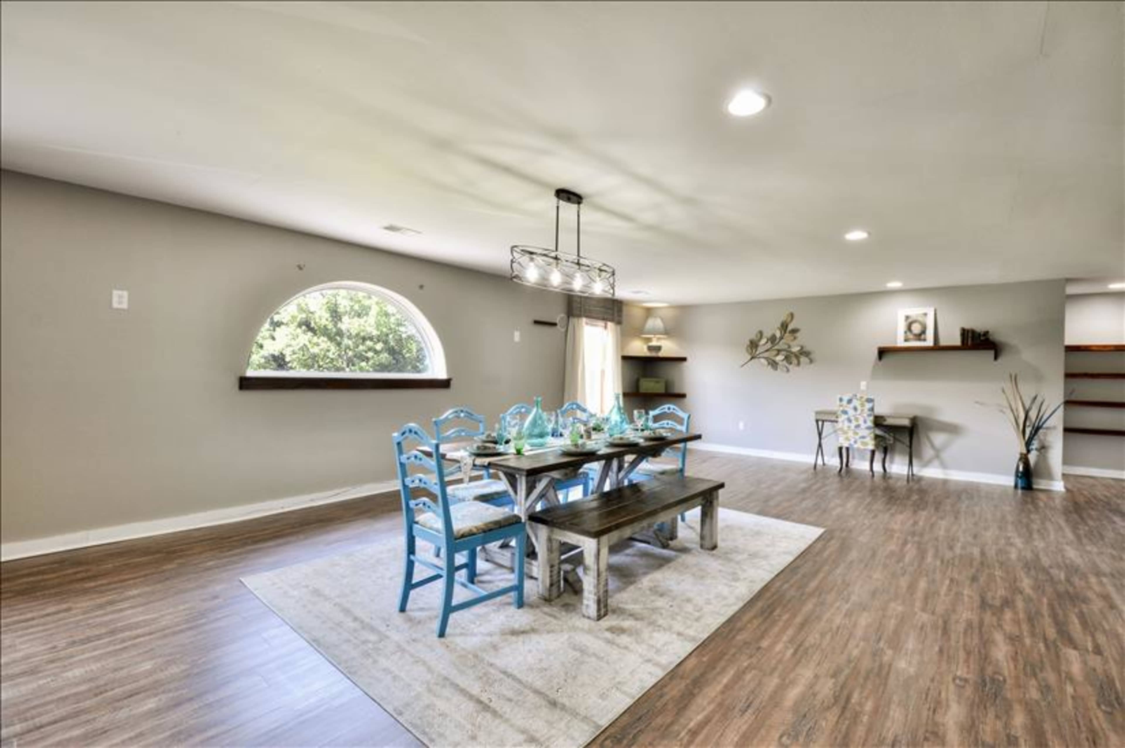A spacious dining area featuring a long wooden table with blue chairs, an oval window, and minimalist decor against light-colored walls.