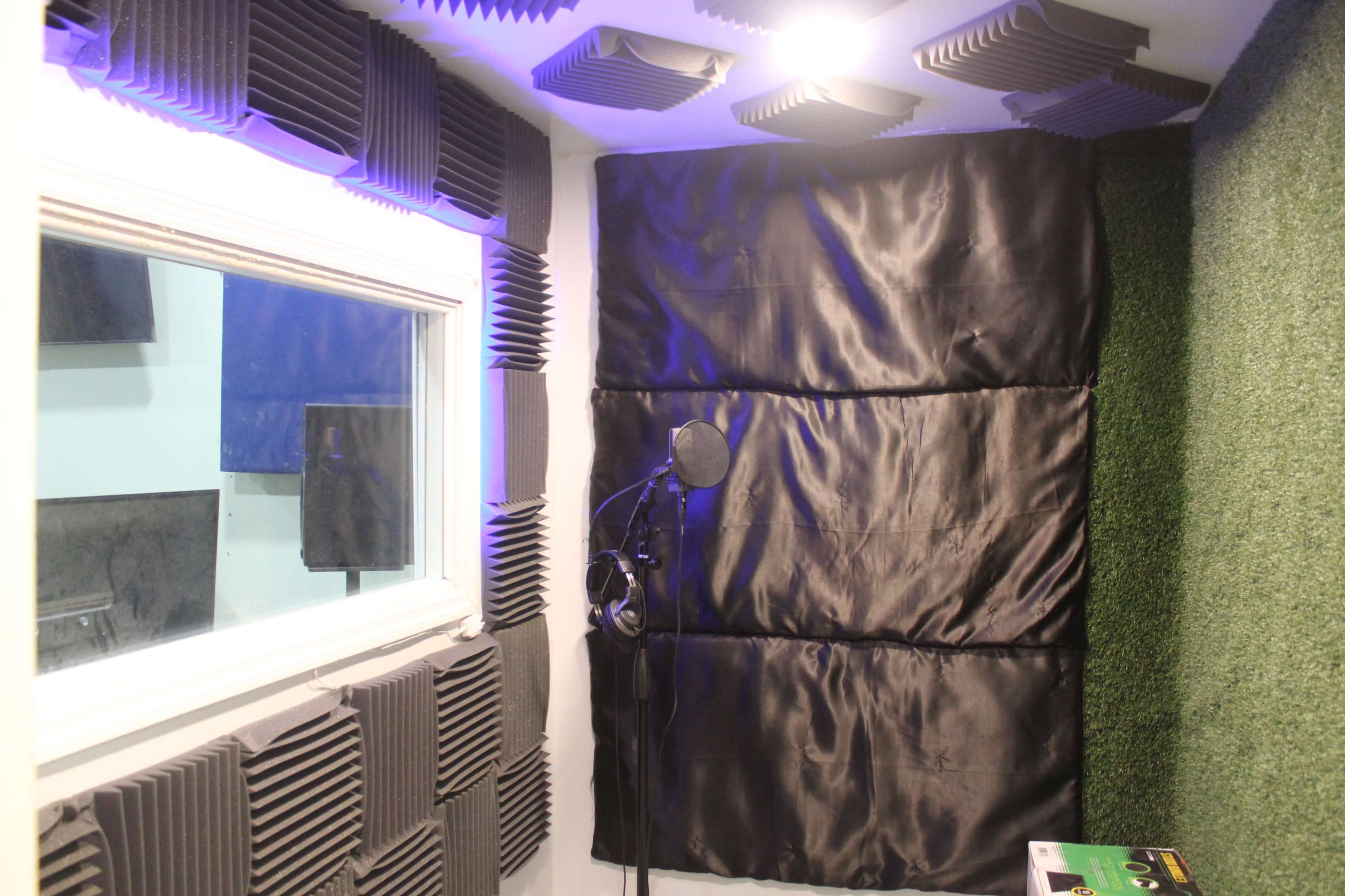 The image shows a soundproof recording booth with acoustic foam panels, a microphone on a stand, and a window for visual access.
