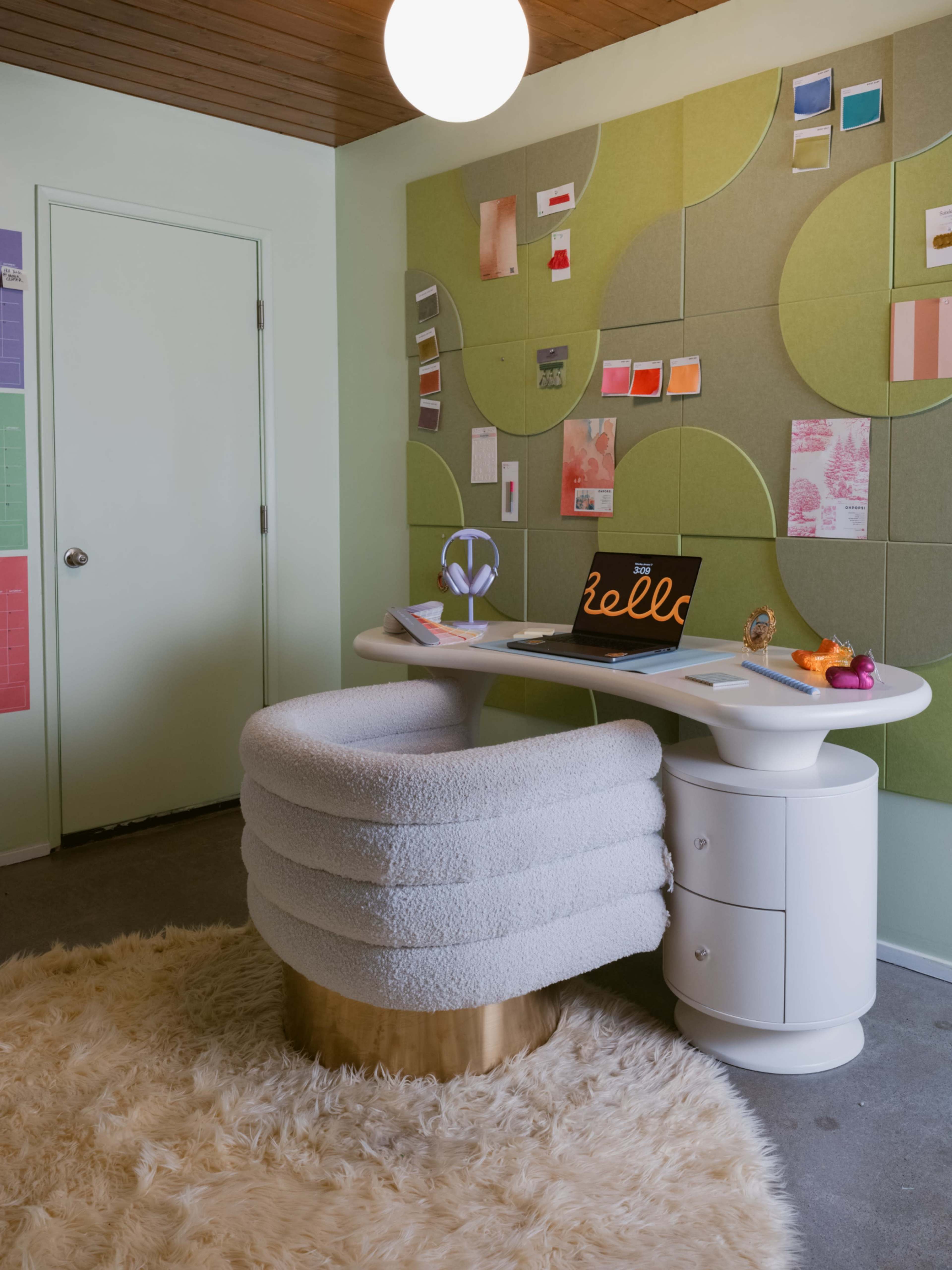 A modern workspace features a curved, upholstered desk chair, a sleek white desk with a laptop, and a colorful wall display of fabric samples and notes.