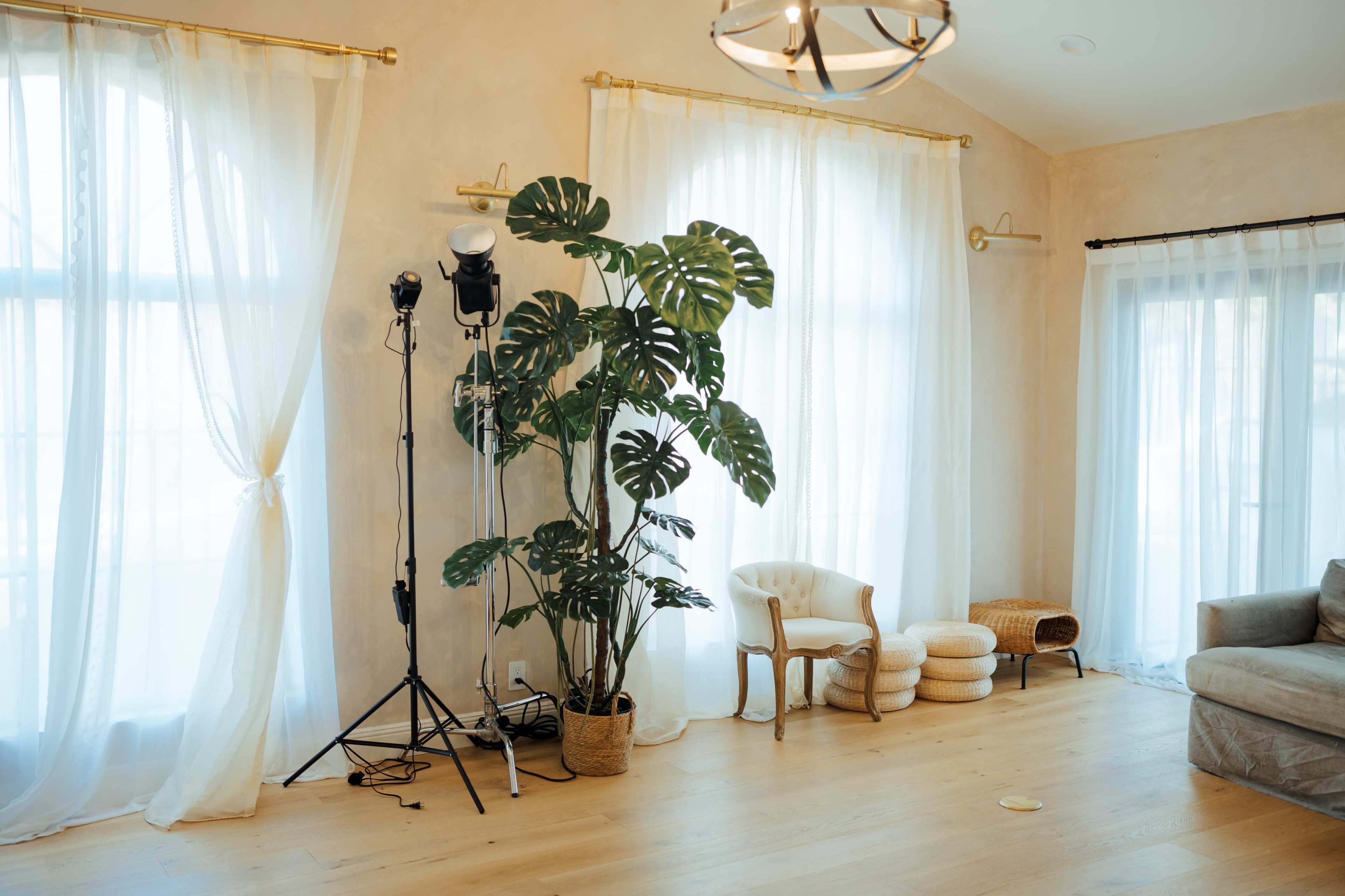 A bright interior room featuring large windows with sheer curtains, a potted plant, a tripod light stand, and a small seating area.