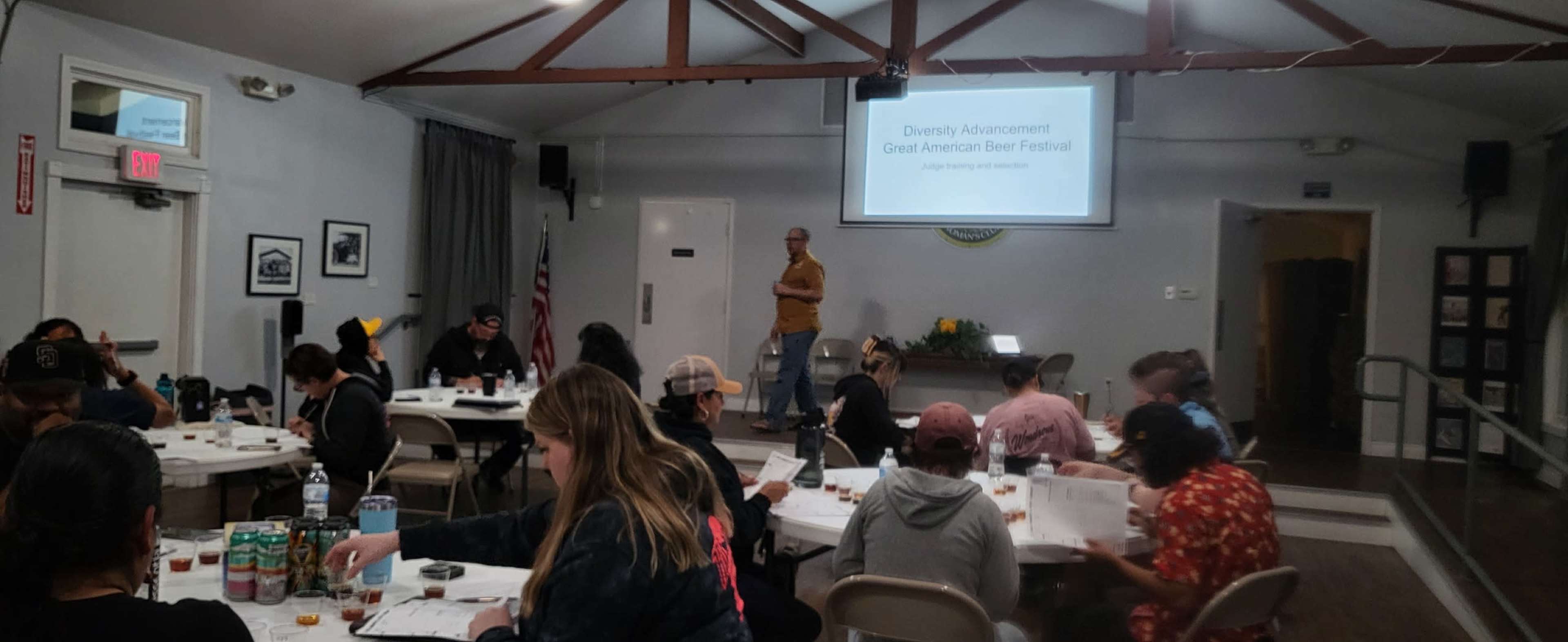 A group of people sit at tables in a room, listening to a speaker presenting a slide titled "Diversity Advancement: Great American Beer Festival."