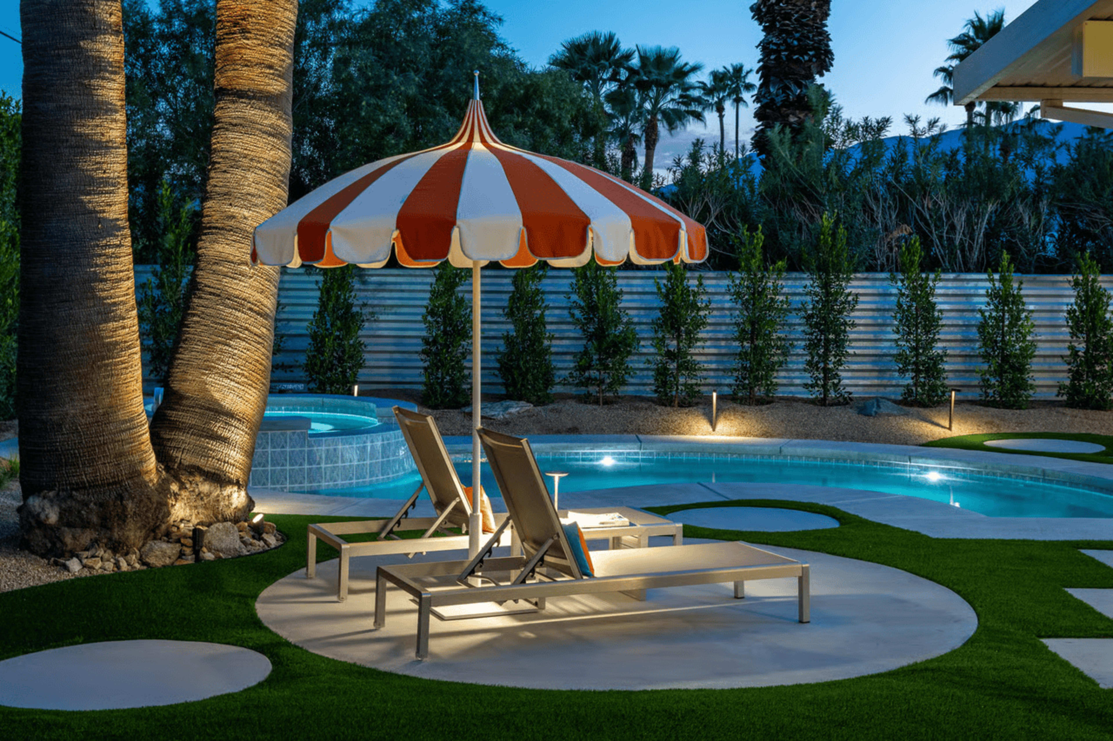The image shows a cozy outdoor pool area featuring a striped umbrella, two lounge chairs, and a well-maintained lawn under a twilight sky.