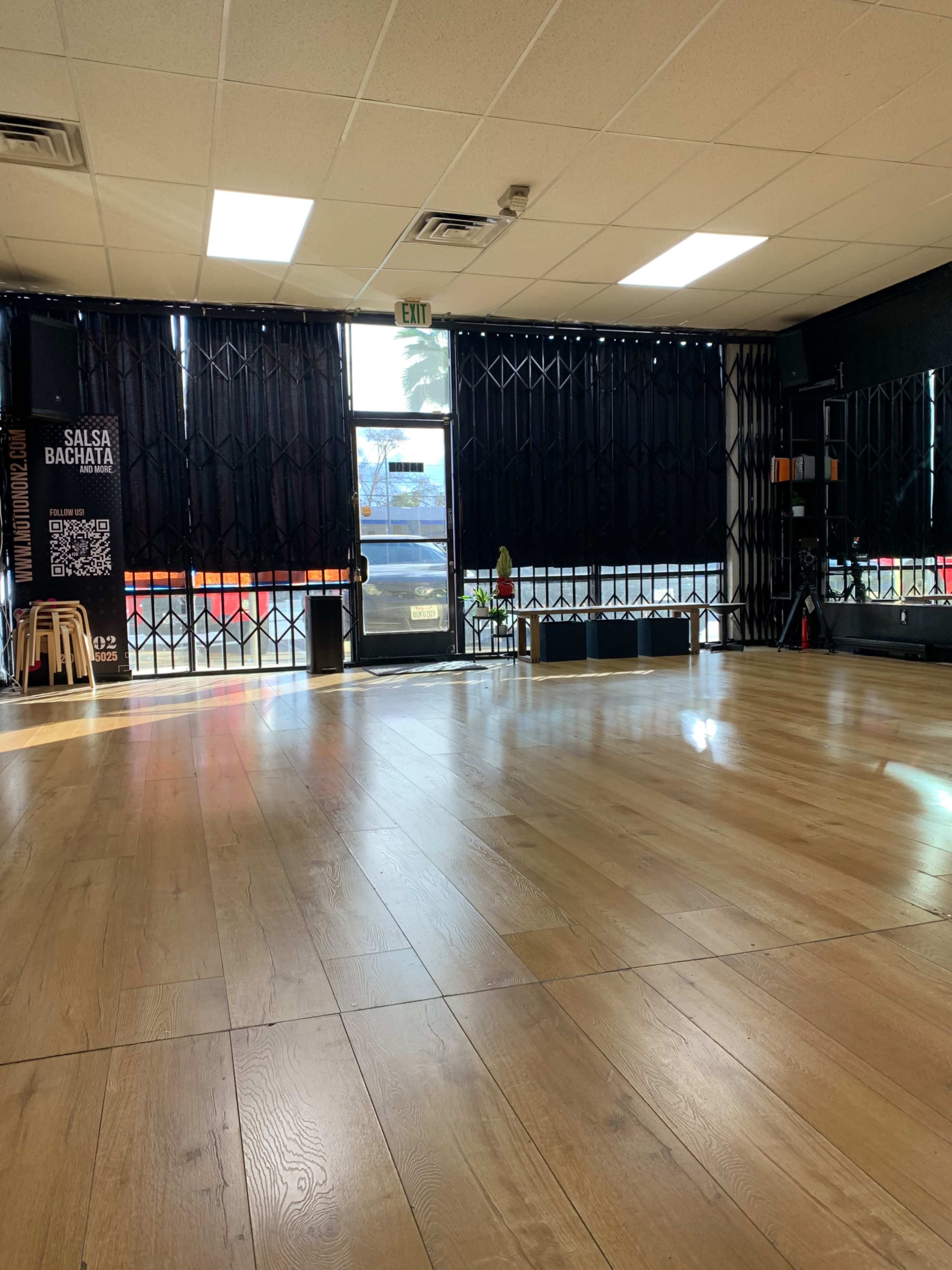 The image shows a spacious dance studio with wooden flooring, a large window covered by black curtains, and a visible entrance with a security gate.
