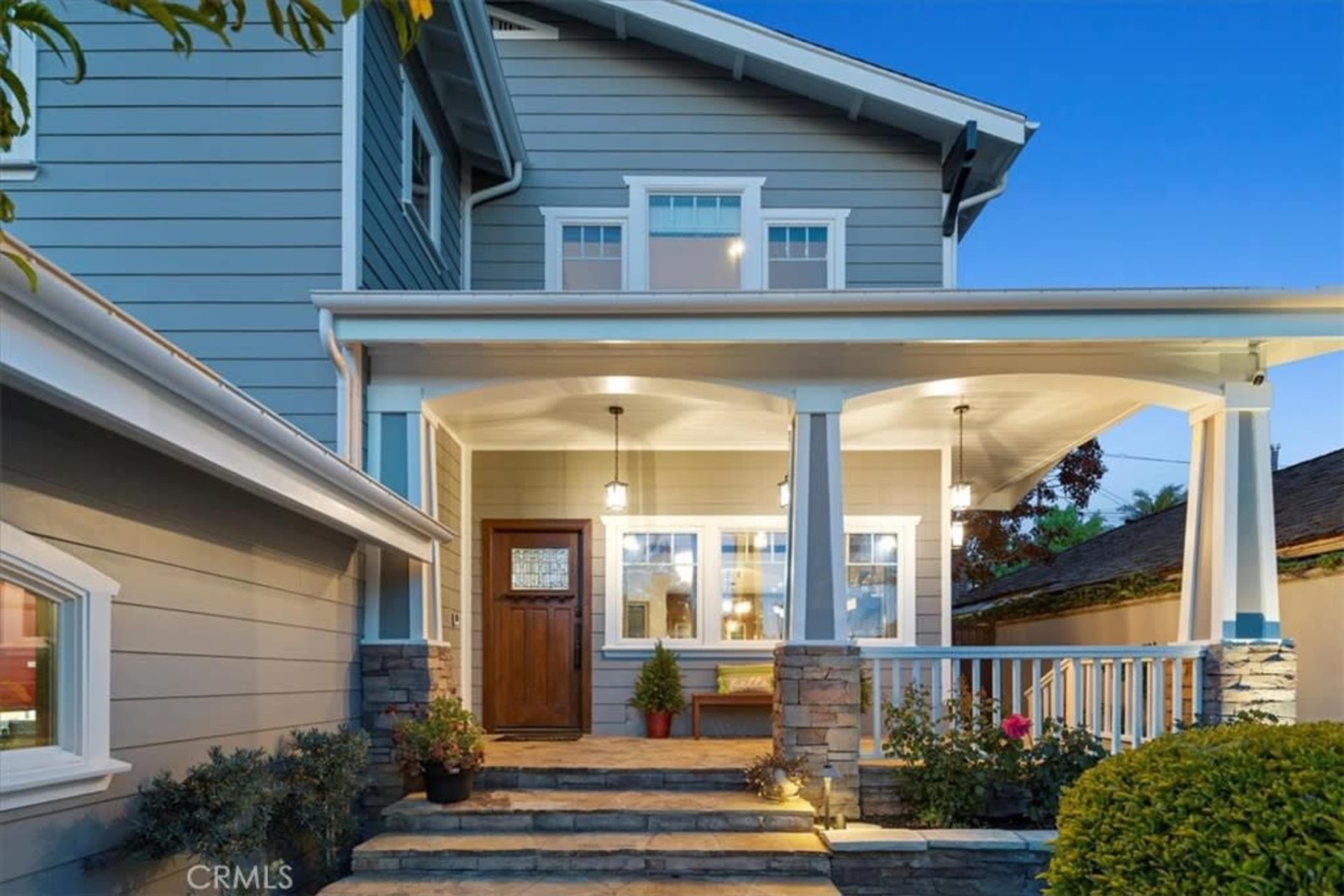 A two-story house with a front porch featuring wooden pillars, stone accents, and potted plants, set against a twilight sky.