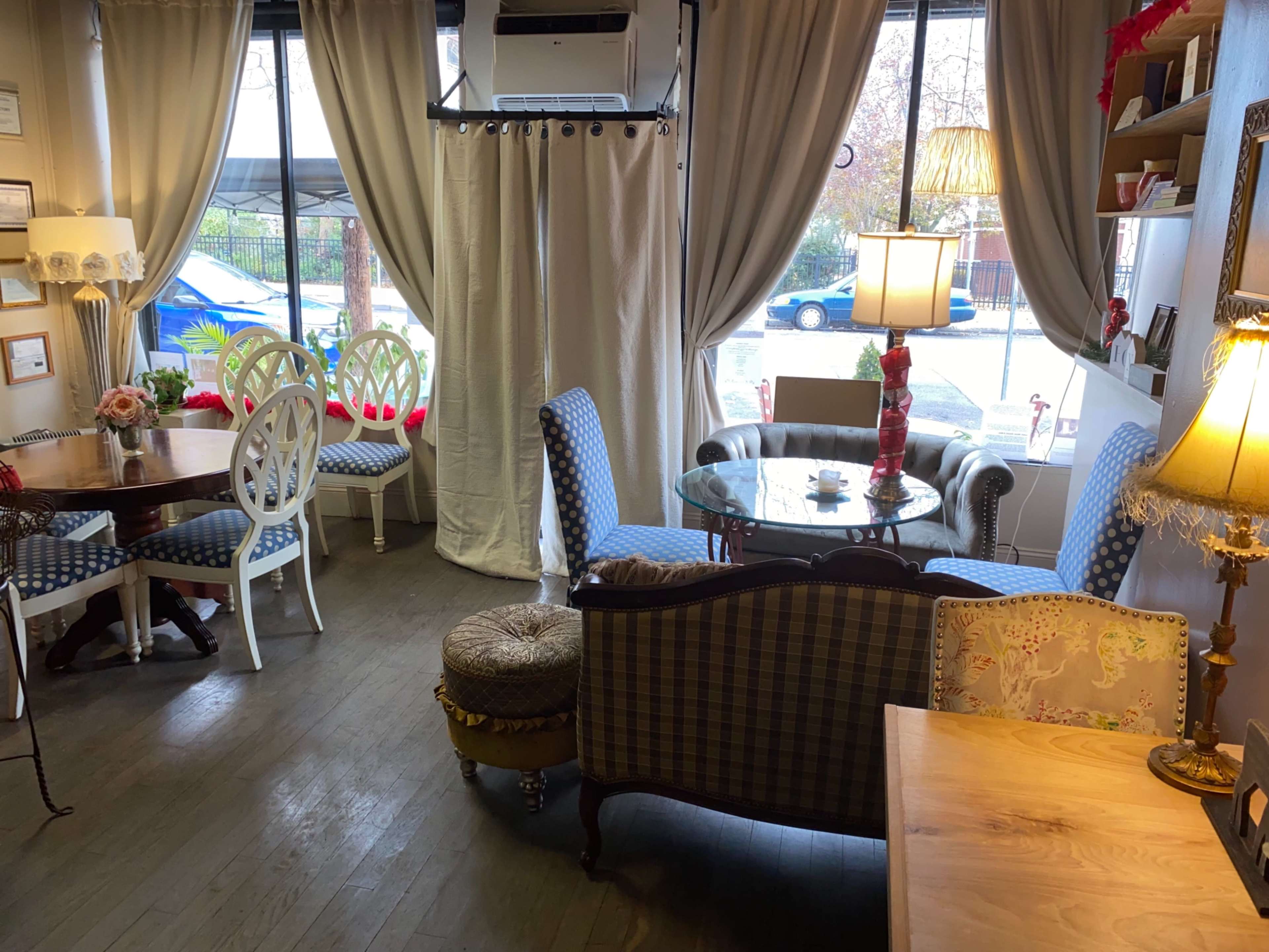 A cozy café interior with a mix of vintage furniture, including a round glass table, patterned chairs, and a small dining table, set against large windows overlooking the street outside.