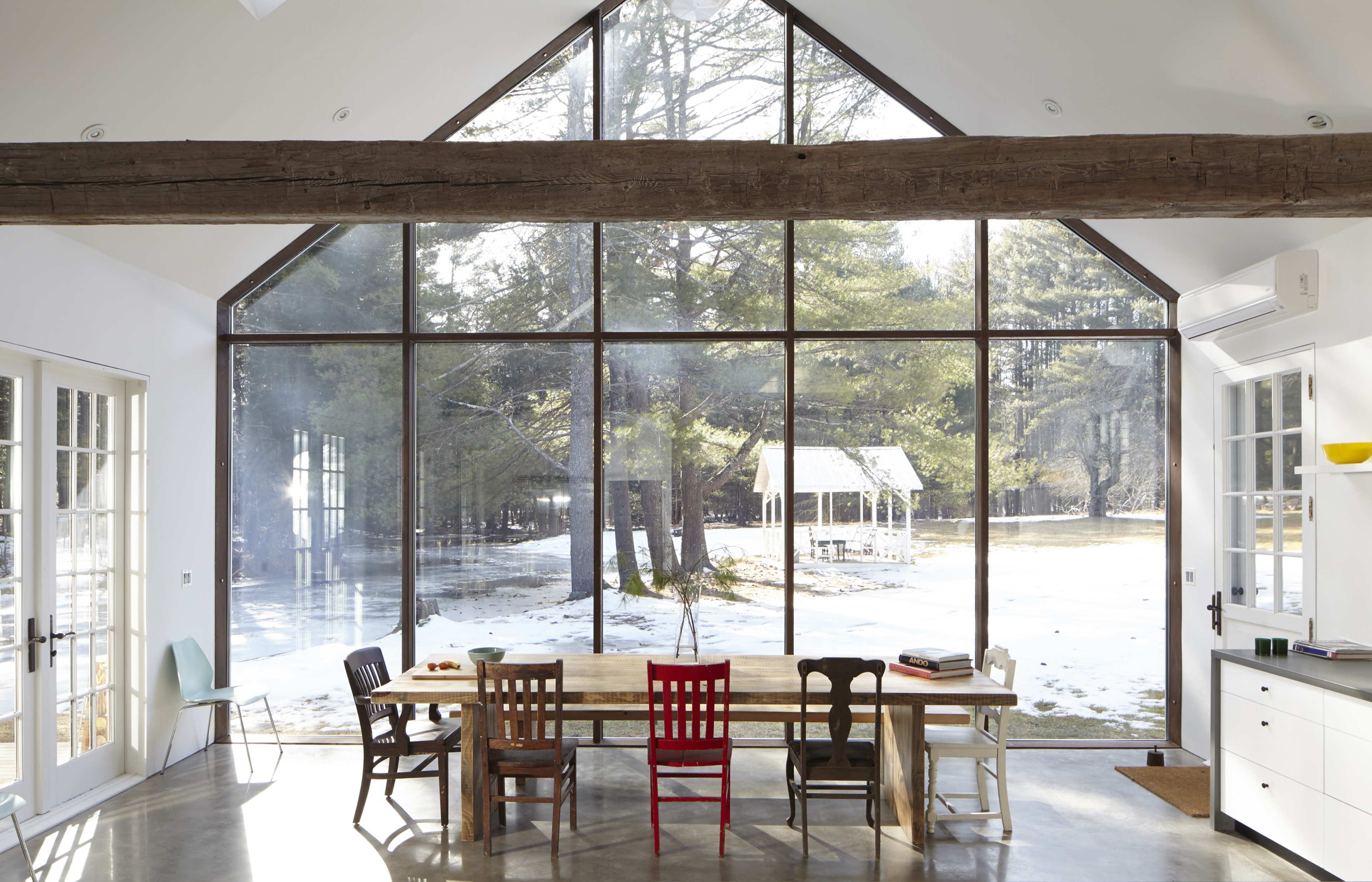 A modern interior features a large dining table surrounded by colorful chairs, with expansive glass walls showcasing a wooded landscape.
