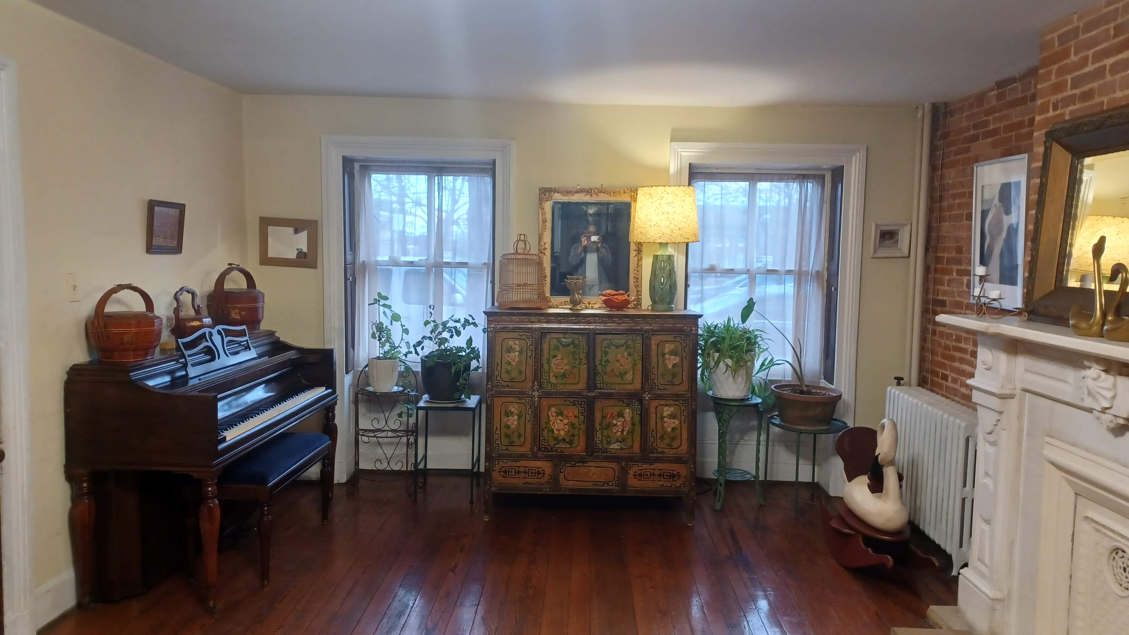 The scene features a living room with a wooden piano, an ornate cabinet, various plants, and a lamp illuminating the space near two windows.