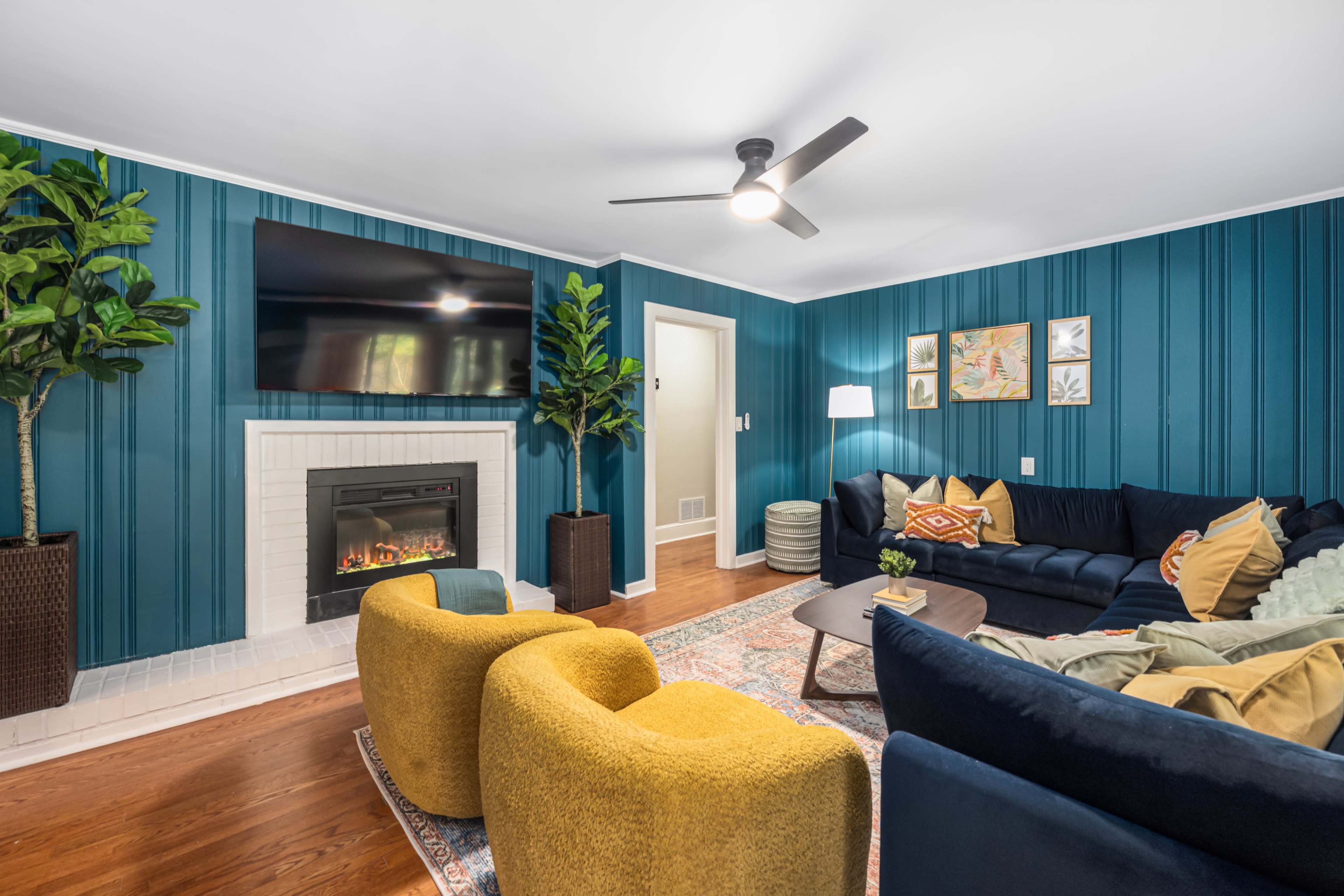 The image shows a living room with teal walls, a fireplace, a television mounted on the wall, and cozy furnishings including a dark blue sofa and two yellow accent chairs.