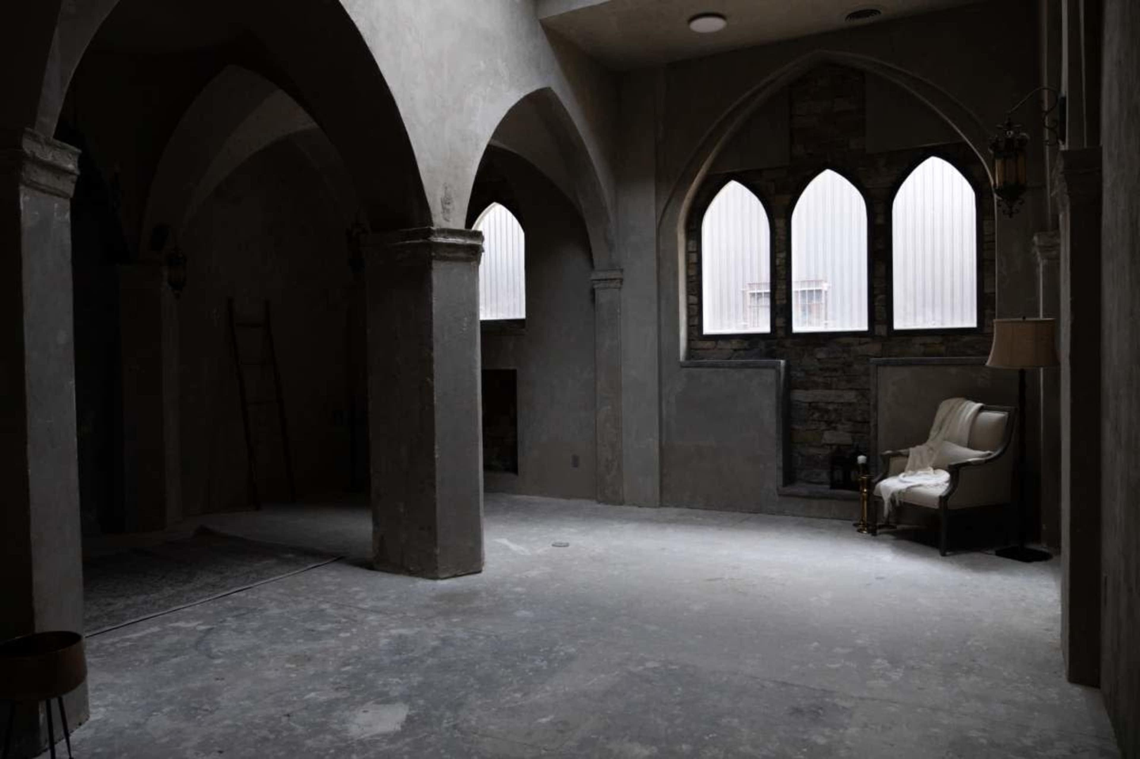 The image shows a dimly lit interior of a room featuring arched columns, stone walls, and a single armchair near large windows.