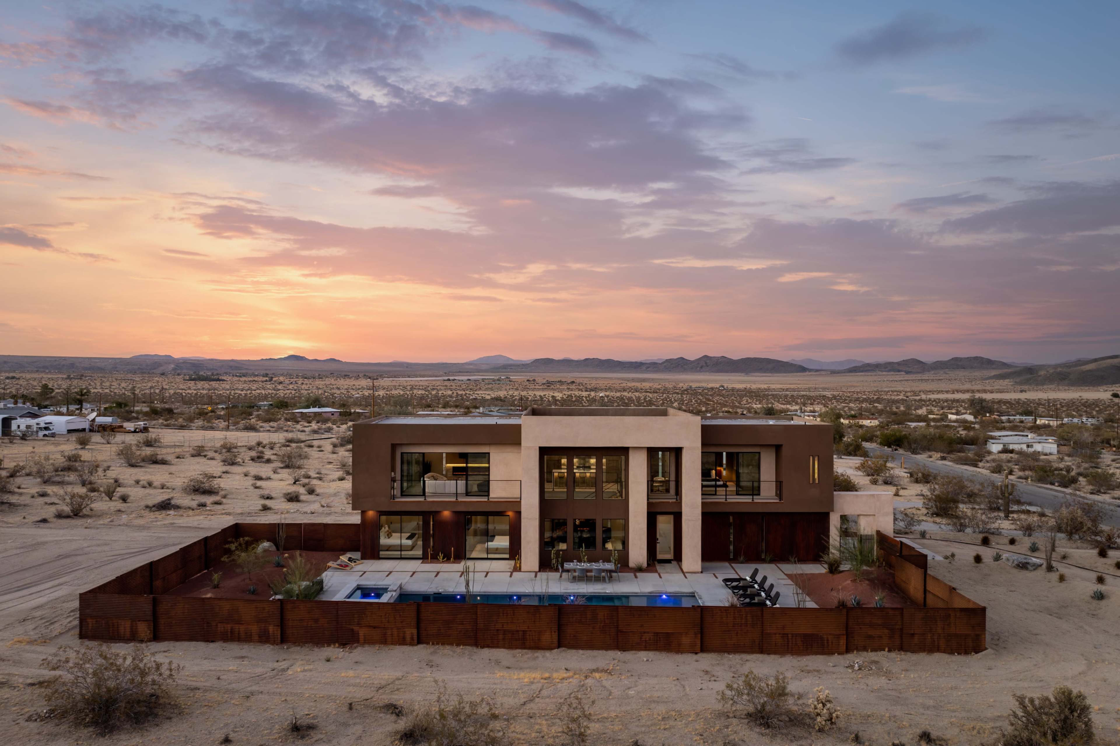 A modern two-story house with a pool is surrounded by desert landscape and mountains under a colorful sunset.