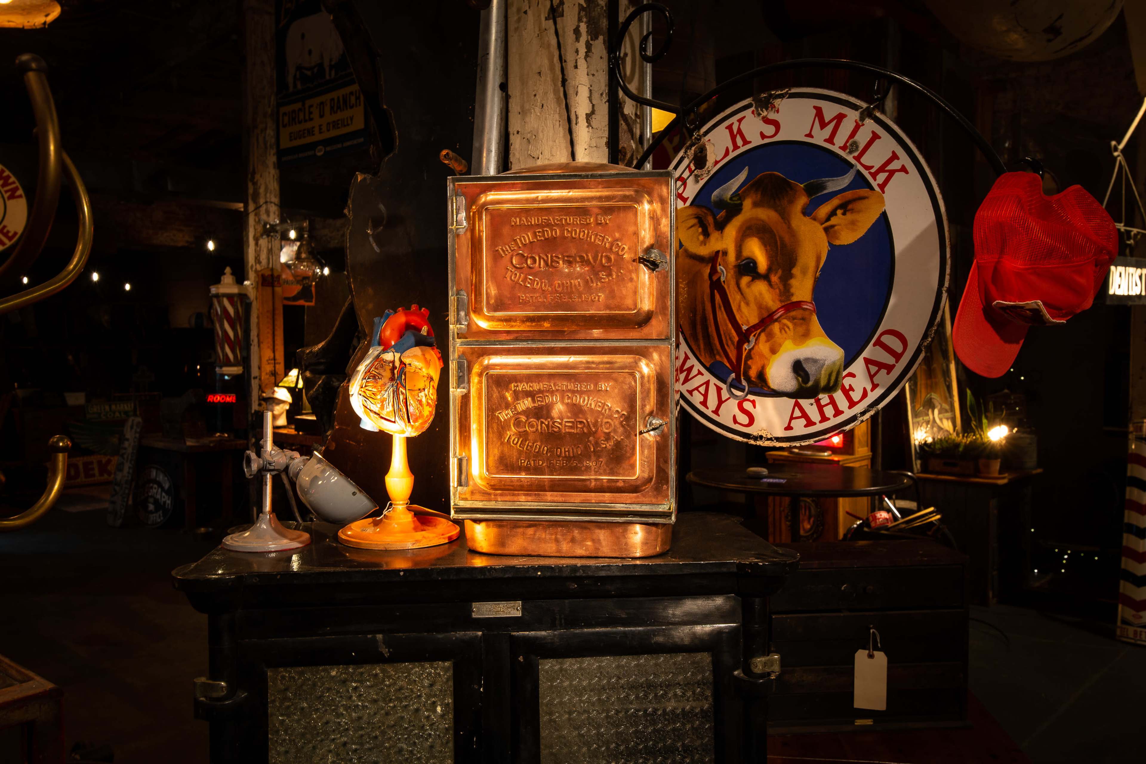 The image shows a vintage display featuring a pair of copper canisters, a small lamp, and a sign depicting a cow, all arranged on a dark surface with various nostalgic items around them.
