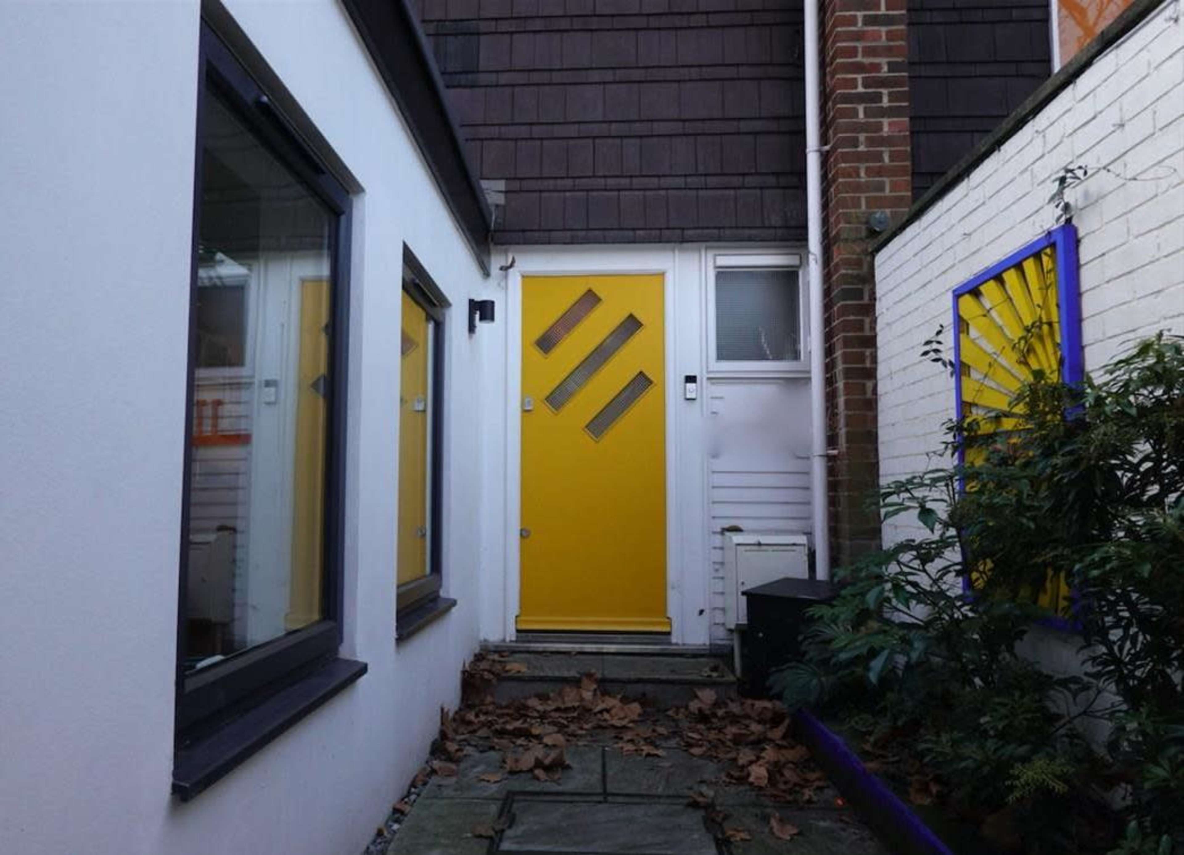 A narrow pathway leads to a bright yellow door flanked by white walls and surrounded by scattered leaves.