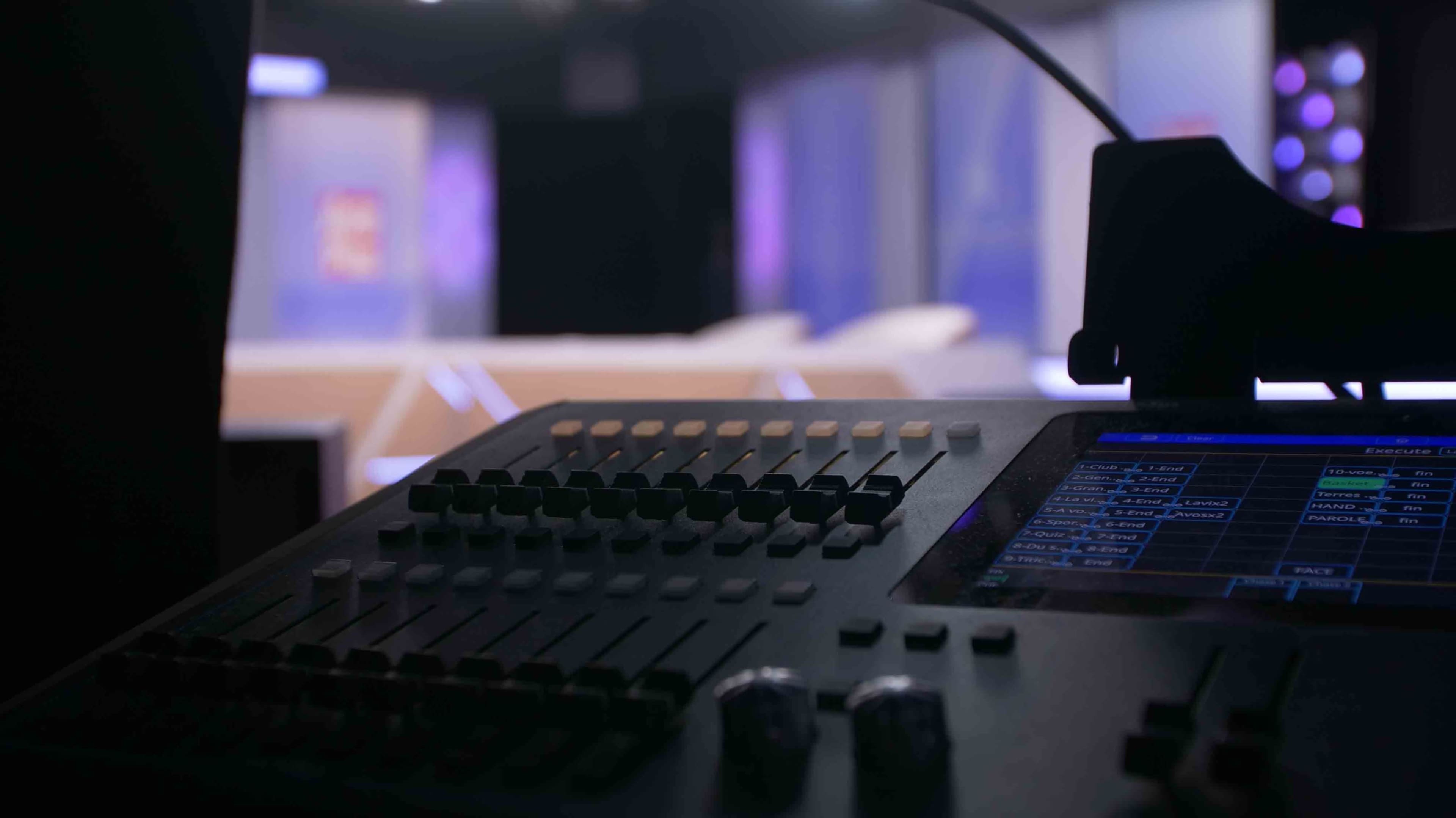 A dimly lit control panel is shown in the foreground with a blurred television studio set in the background.