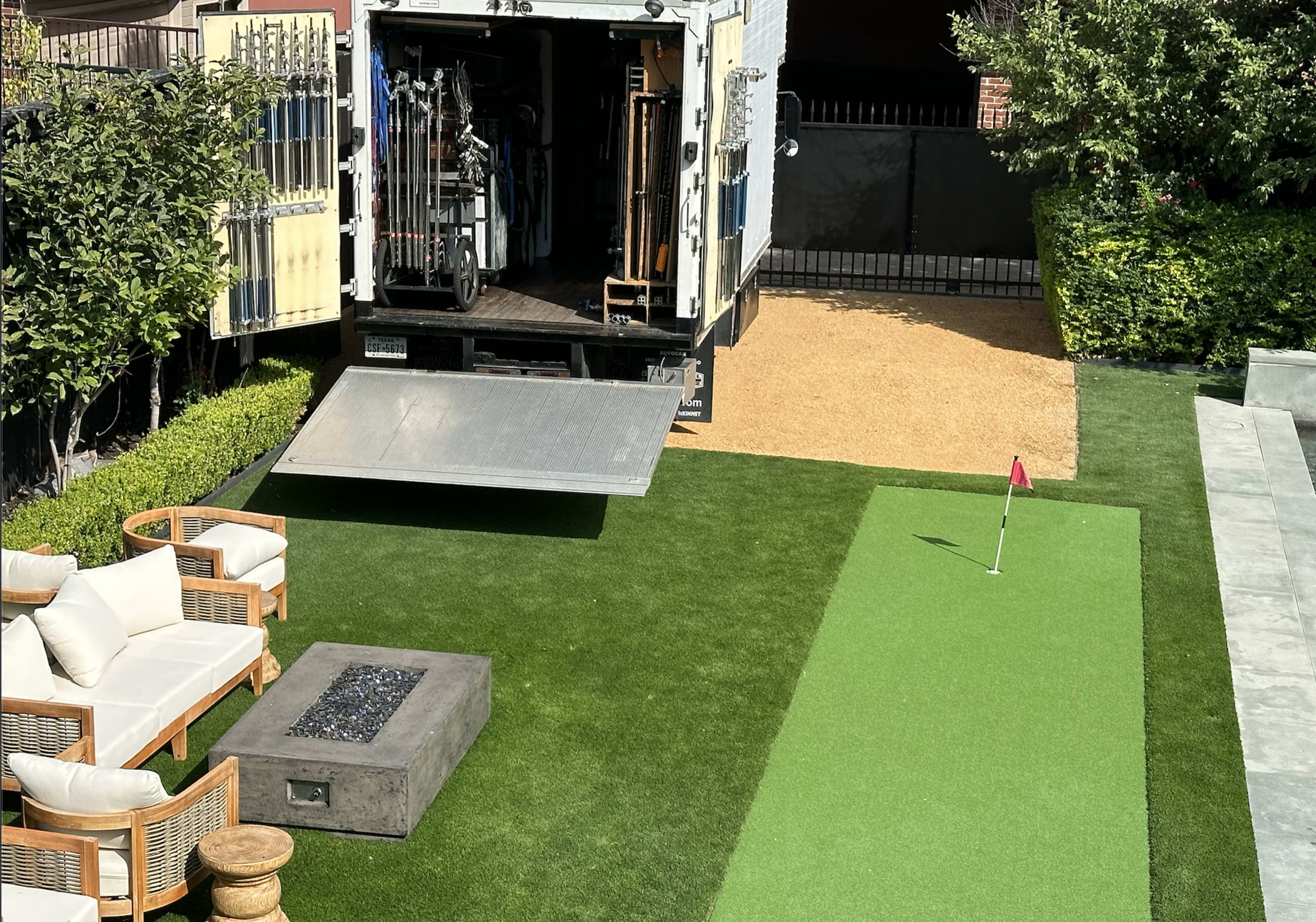 A delivery truck is parked in a yard with a putting green and outdoor seating area nearby.