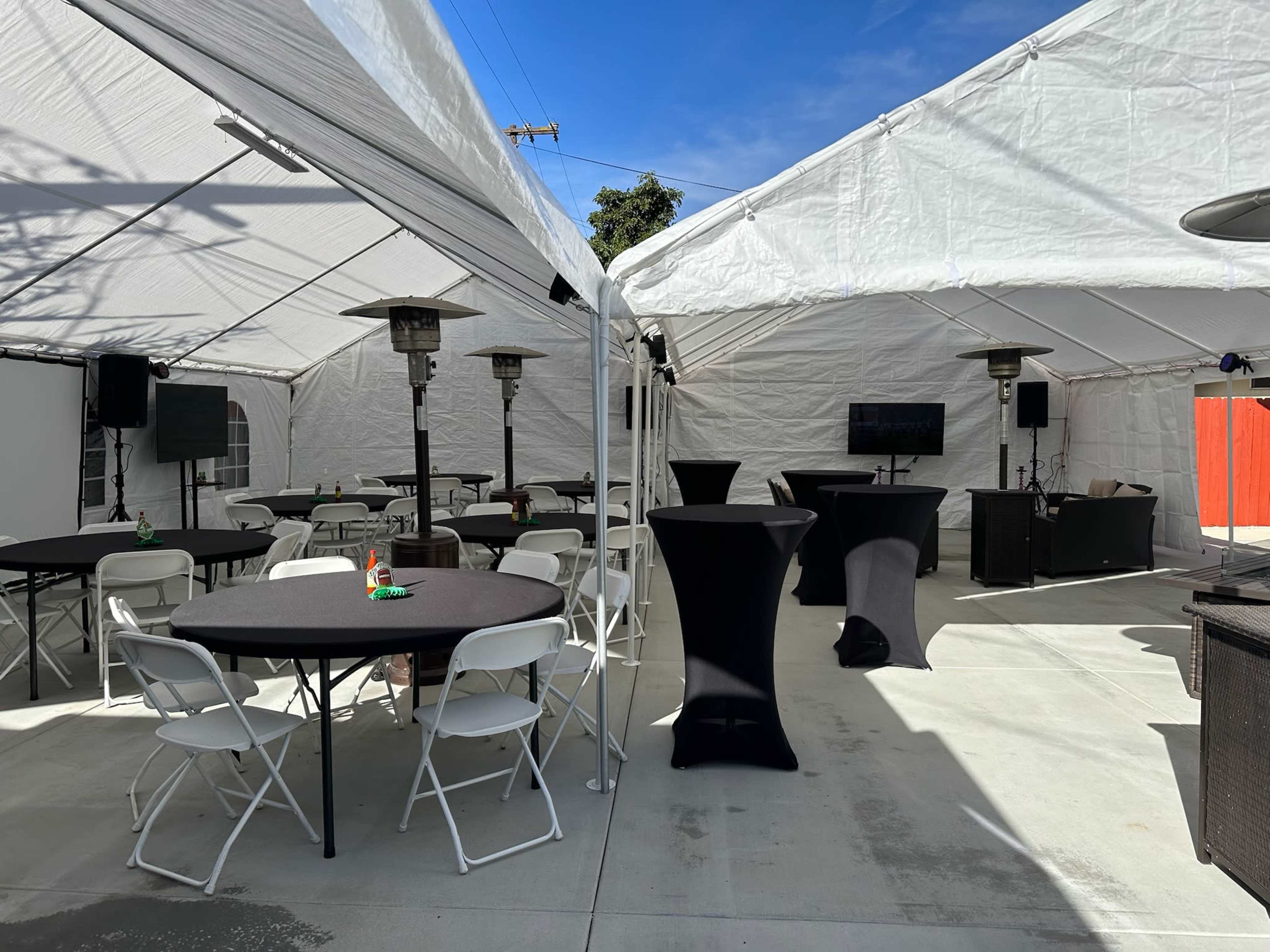 The image shows a setup of two large white tents with tables and chairs arranged inside, along with outdoor heaters and screens.